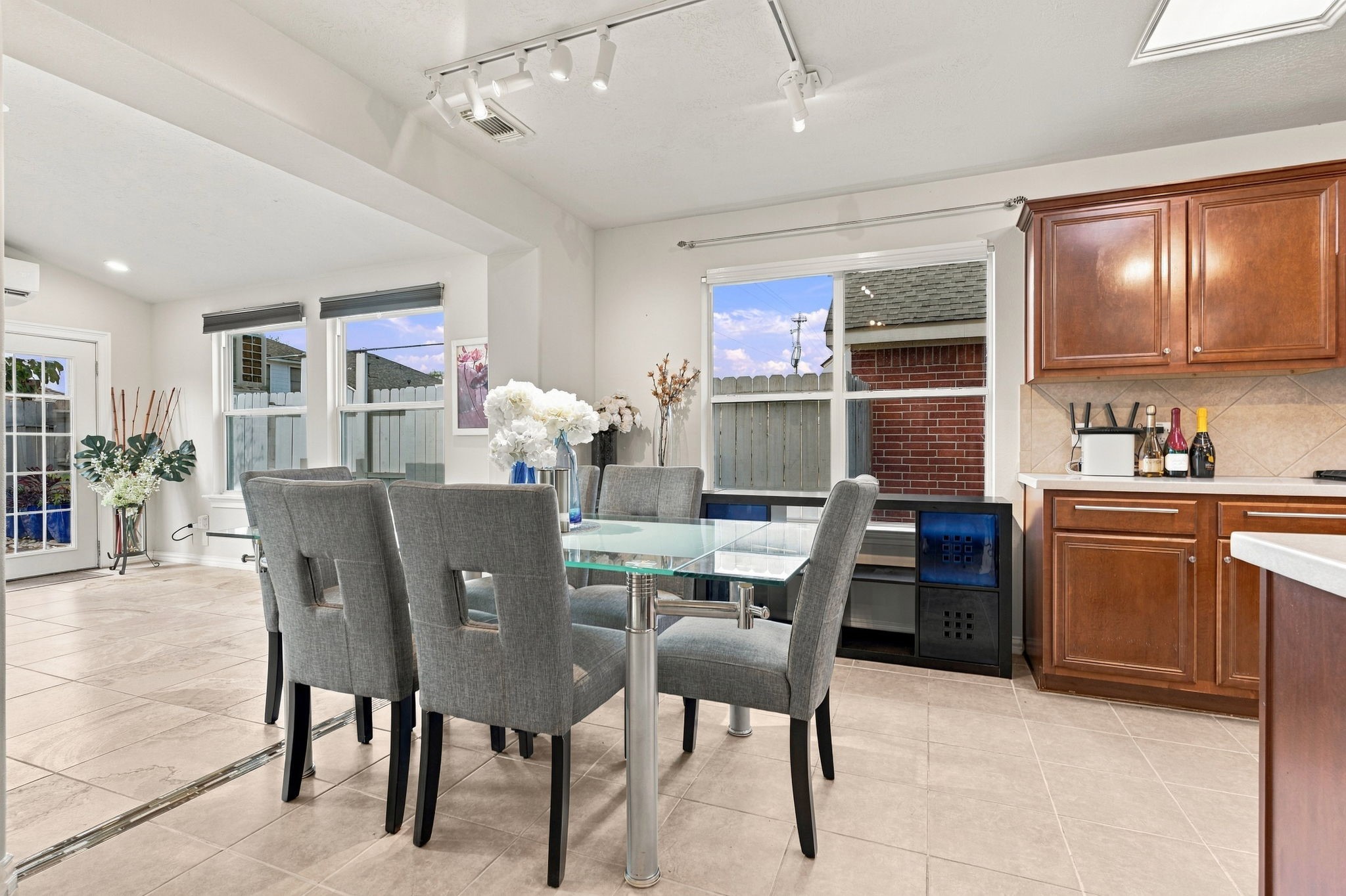 Bright and inviting dining area with modern glass table, elegant gray chairs, and ample natural light, seamlessly connected to the kitchen with rich wooden cabinetry.