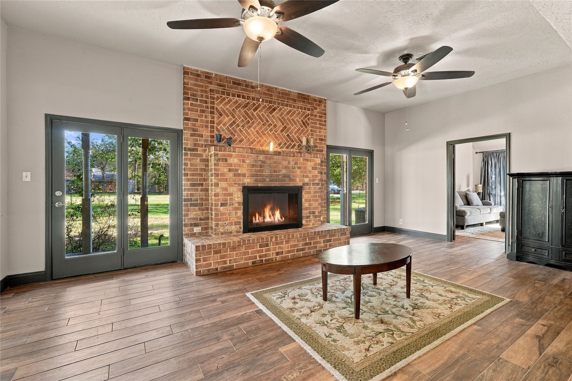 Spacious living room featuring a striking brick fireplace, dual ceiling fans, and glass doors providing views of the surrounding property.
