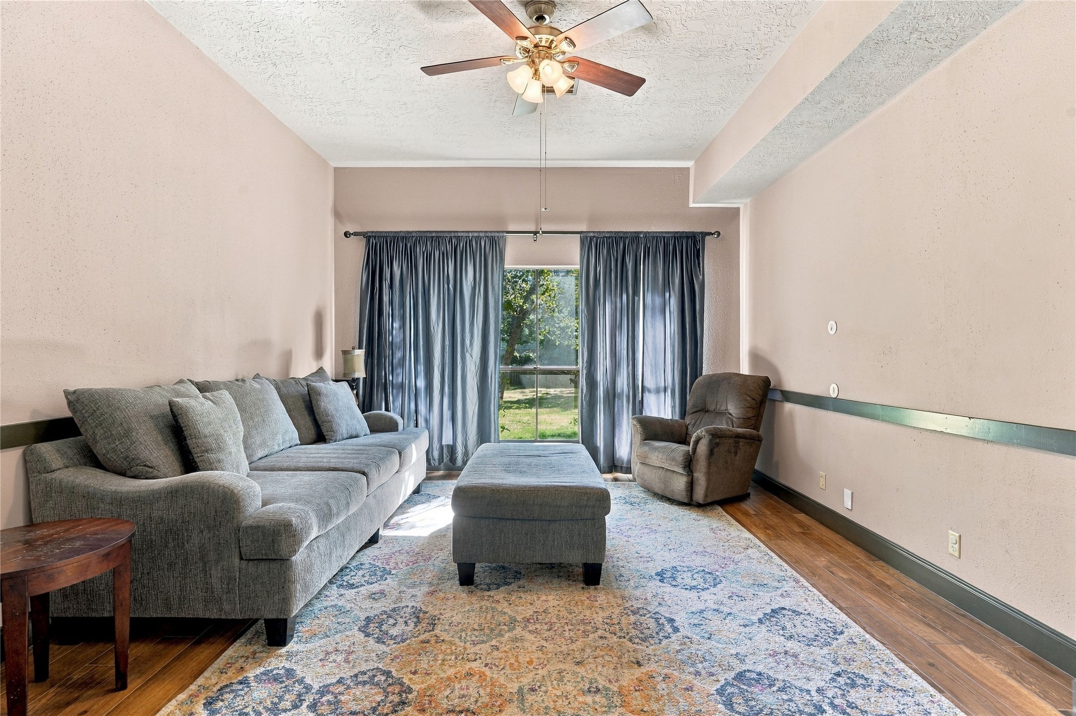 Comfortable family room featuring wood-look flooring, ceiling fan, and large window overlooking the yard.