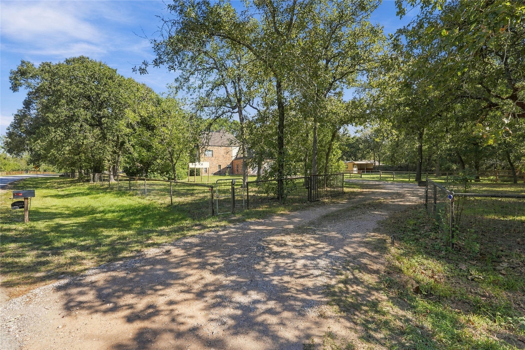 Gated property entrance with a gravel driveway surrounded by mature trees and natural landscaping.