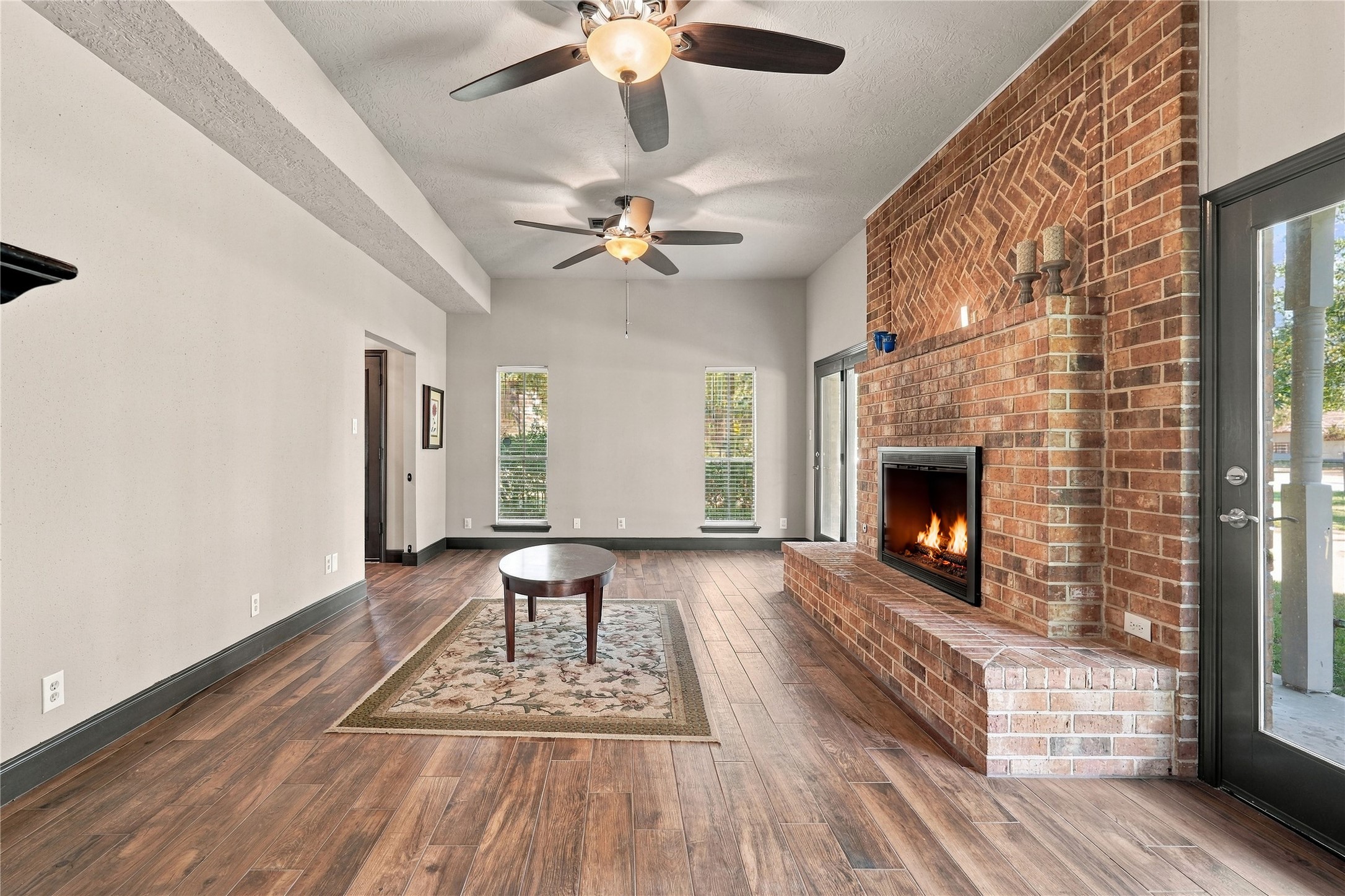 Spacious living room featuring a stunning floor-to-ceiling brick fireplace, wood-look flooring, abundant natural light, and French doors providing access to the covered porch.