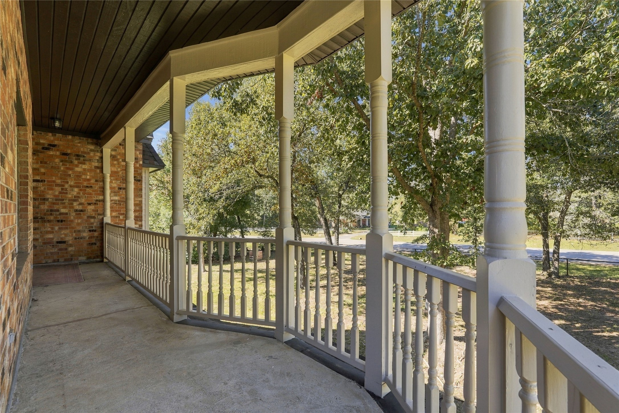Expansive covered balcony featuring decorative columns, railing, and peaceful views of the mature trees and surrounding property.