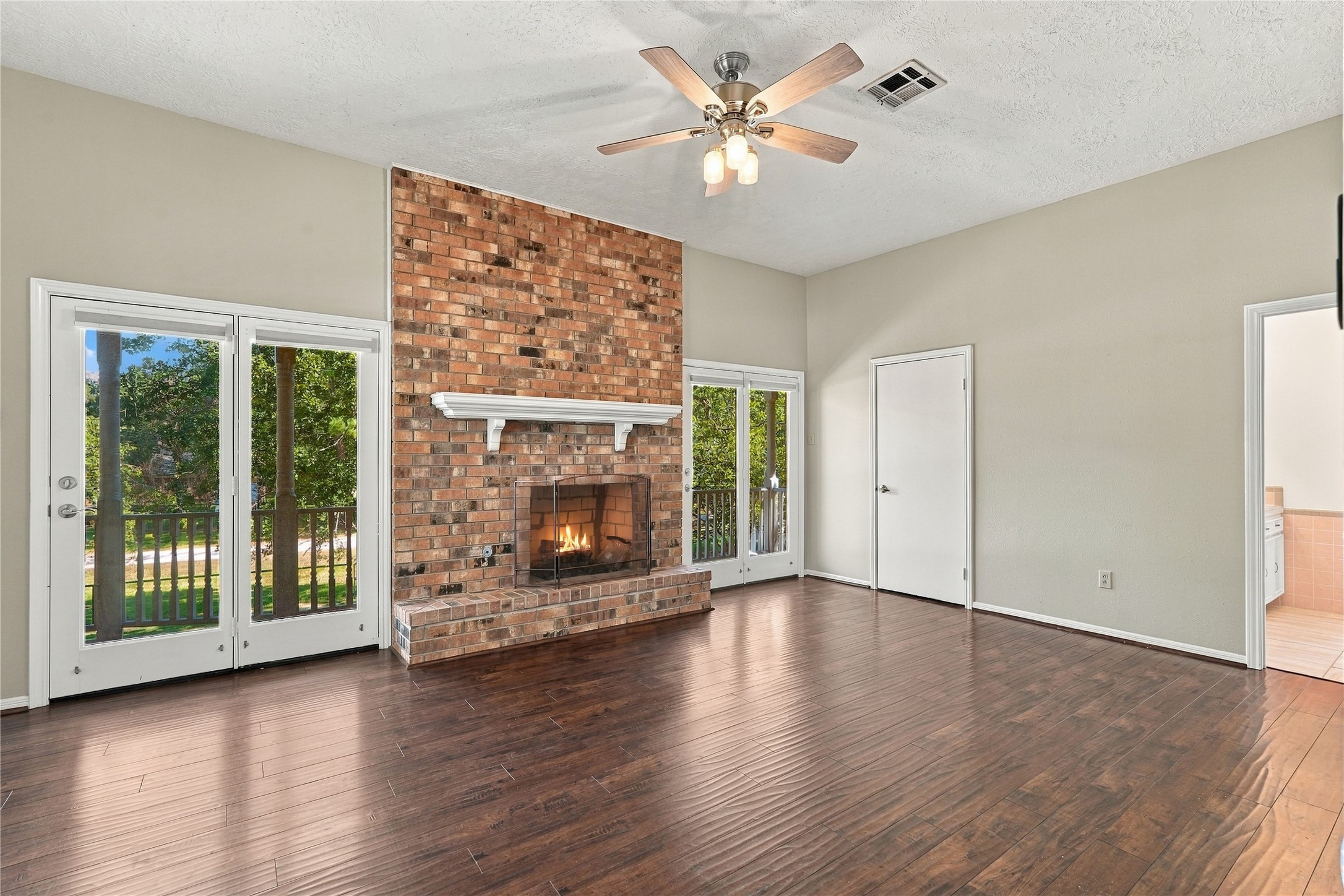 Spacious primary bedroom featuring a floor-to-ceiling brick fireplace, wood flooring, ceiling fan, and French doors opening to the covered balcony.