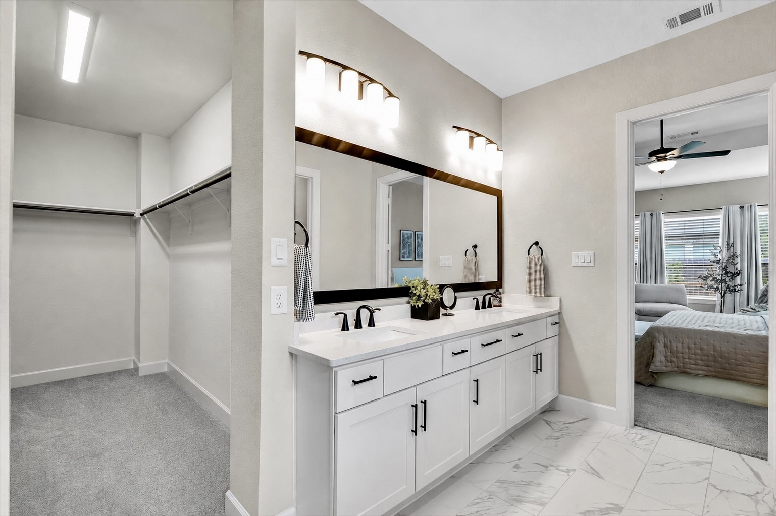 Bright, modern bathroom with dual sinks and sleek lighting opens to a spacious walk-in closet and a cozy bedroom with large windows.