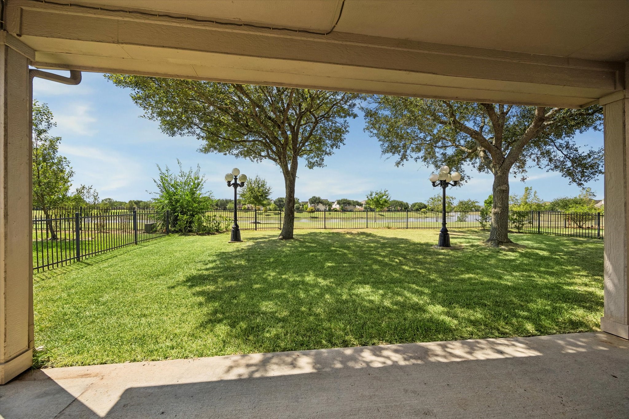 Enjoy the serene views from this covered patio, overlooking a spacious, well-maintained lawn with mature trees and charming lampposts, all enclosed by a classic wrought-iron fence.