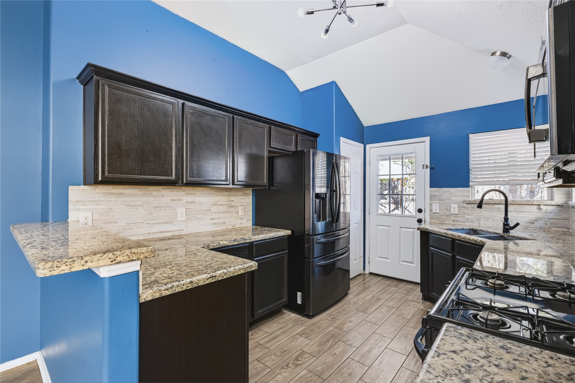 Well-appointed kitchen featuring dark cabinetry, gleaming granite countertops, a gas cooktop, sleek black appliances, and a stylish tiled backsplash with direct access to the backyard. Well-appointed kitchen featuring dark cabinetry, gleaming granite countertops, a gas cooktop, sleek black appliances, and a stylish tiled backsplash with direct access to the backyard.