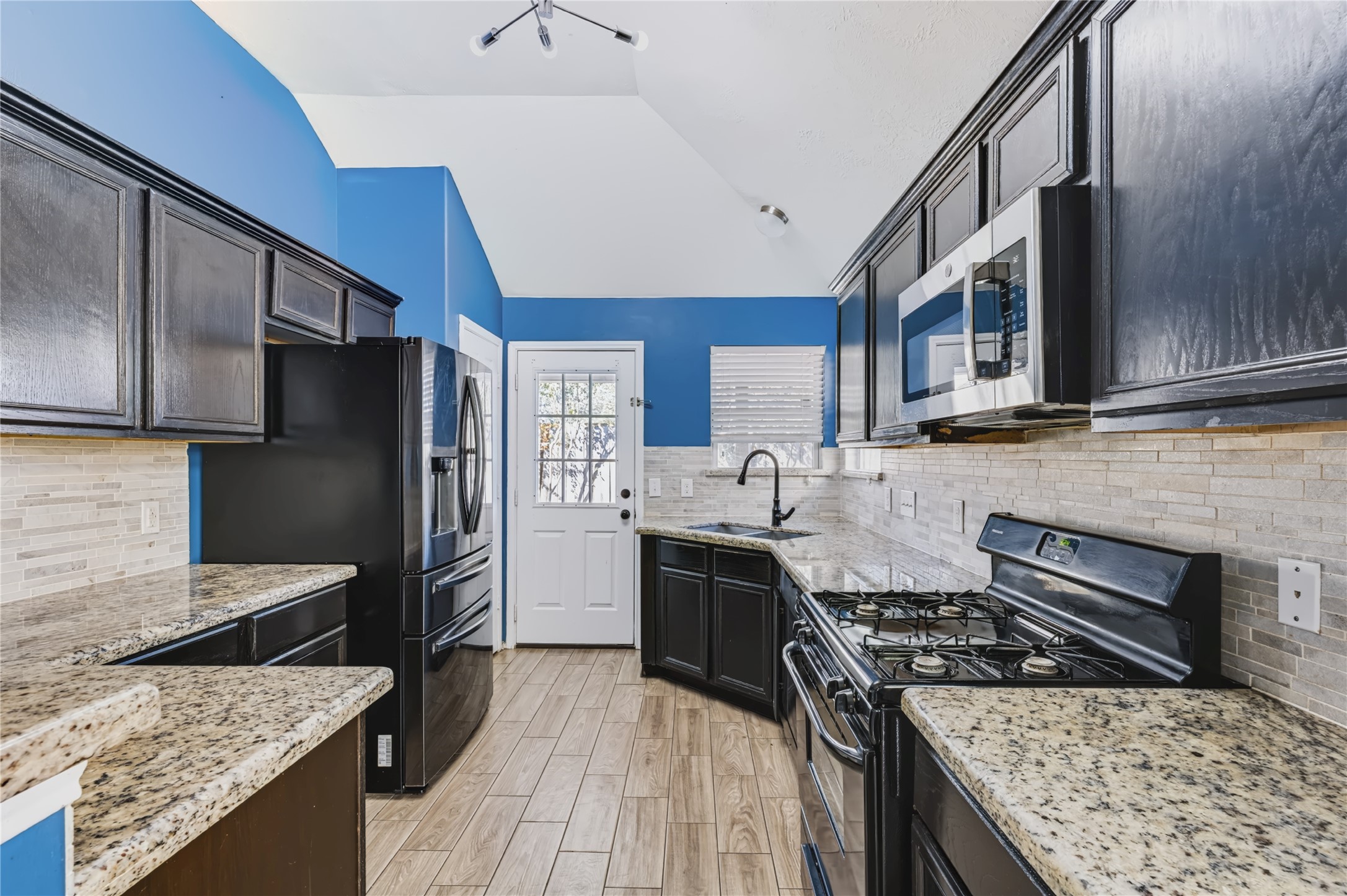 Well-appointed kitchen featuring dark cabinetry, gleaming granite countertops, a gas cooktop, sleek black appliances, and a stylish tiled backsplash with direct access to the backyard. Well-appointed kitchen featuring dark cabinetry, gleaming granite countertops, a gas cooktop, sleek black appliances, and a stylish tiled backsplash with direct access to the backyard.