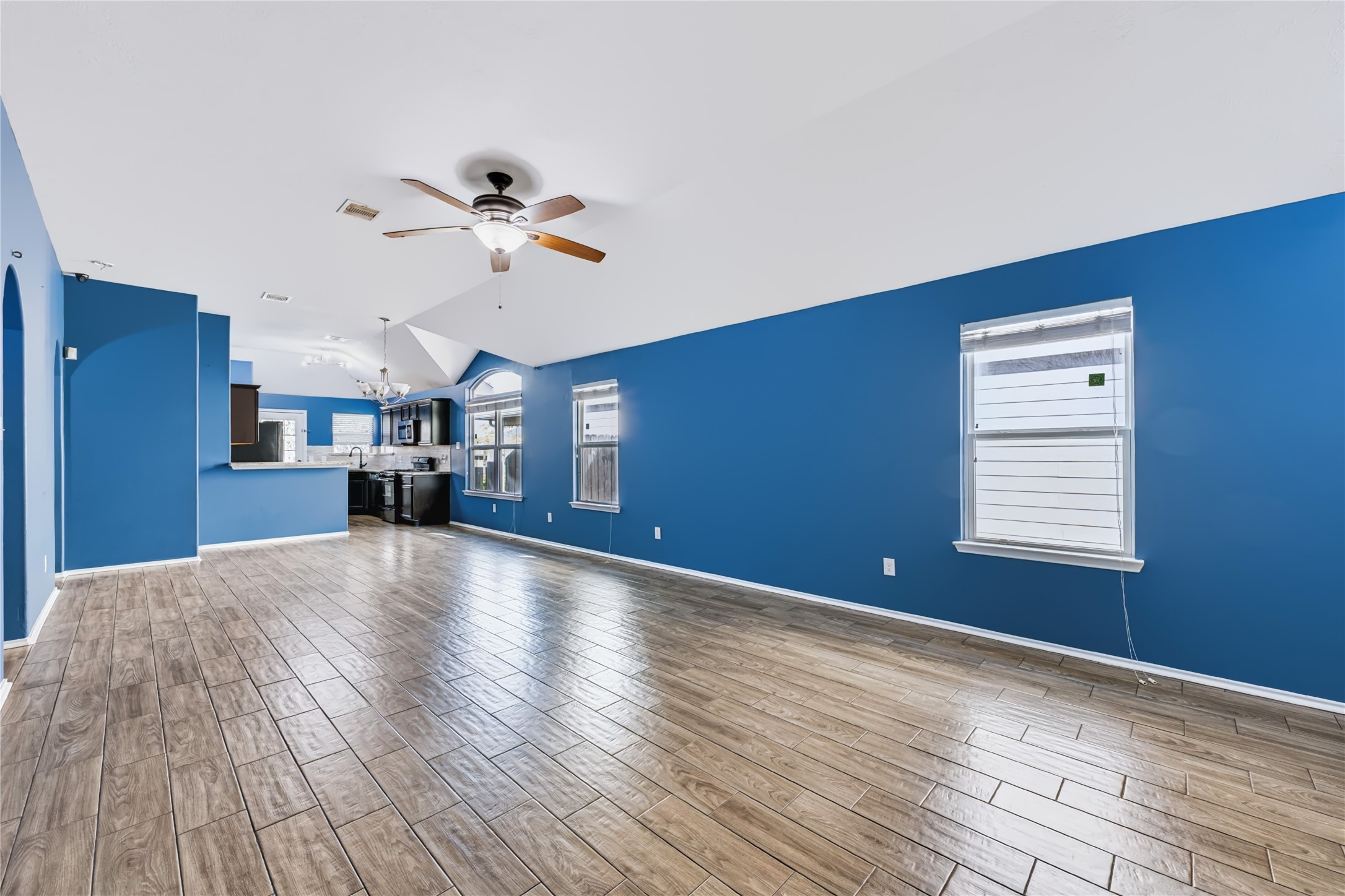 Spacious open-concept living area featuring rich wood-look tile flooring, vaulted ceilings, a modern ceiling fan, and vibrant accent walls that flow seamlessly into the kitchen. Spacious open-concept living area featuring rich wood-look tile flooring, vaulted ceilings, a modern ceiling fan, and vibrant accent walls that flow seamlessly into the kitchen.
