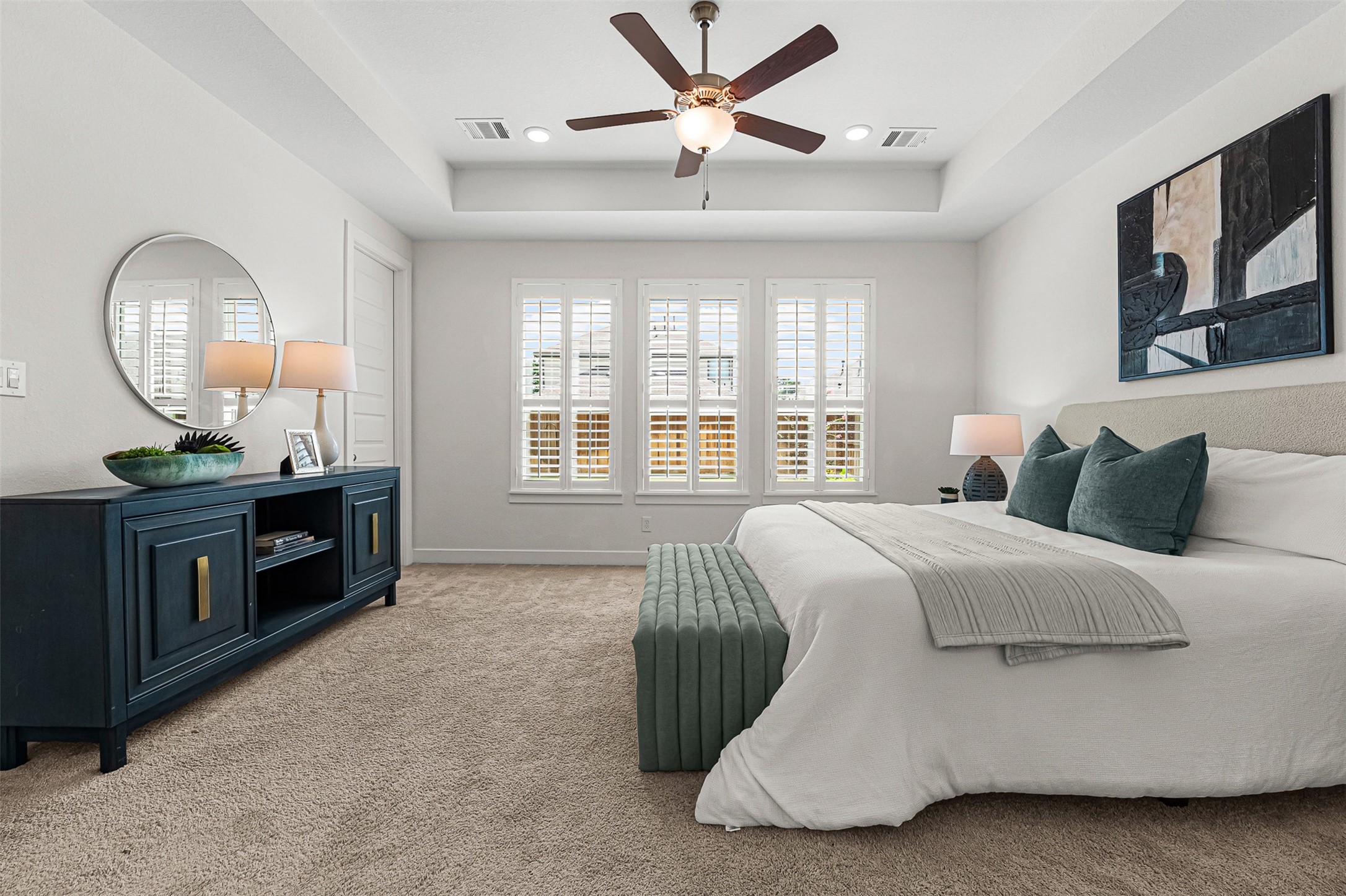 A recessed tray ceiling with a stylish fan elevates the spacious primary suite on the first floor. Planted windows with plantation shutters frame neighborhood views, while soft carpet flooring creates an inviting atmosphere for relaxation.