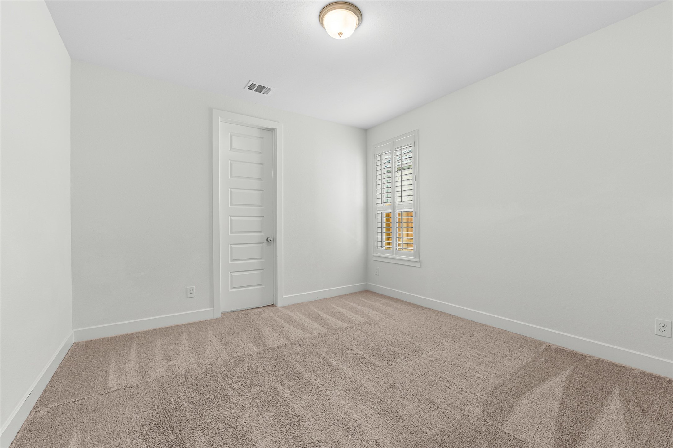 Plush carpet flooring and crisp plantation shutters define this sunlit secondary bedroom, creating a peaceful and versatile space. Crisp white walls and a six-panel interior door complete the bright, neutral design.