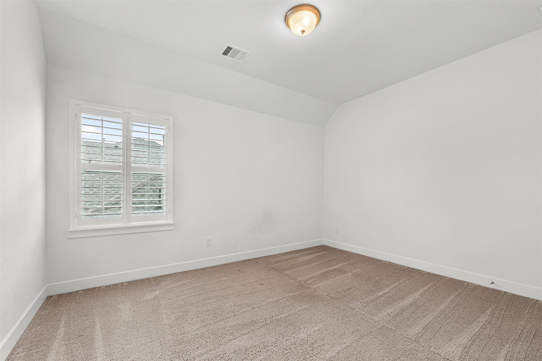 Plush neutral carpeting and classic white plantation shutters define this sunlit secondary bedroom. Sloped ceiling accents and crisp white walls enhance the versatile space.