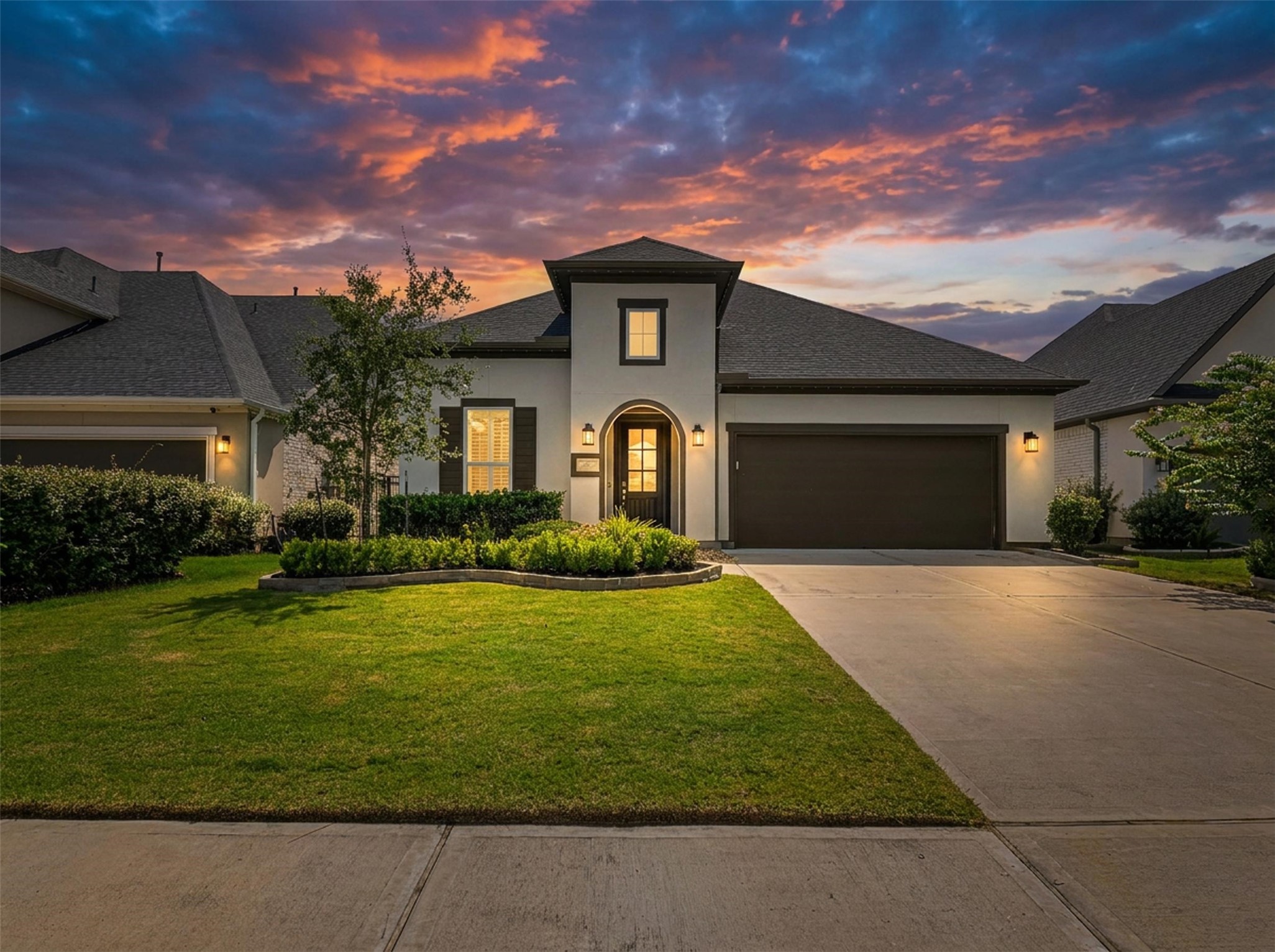A grand two-story elevation showcases sleek stucco, dark accent shutters, stone-bordered landscaping, and an arched entry under sunset skies. An expansive driveway leads to the attached two-car garage, completing the stunning exterior curb appeal of this Houston home.