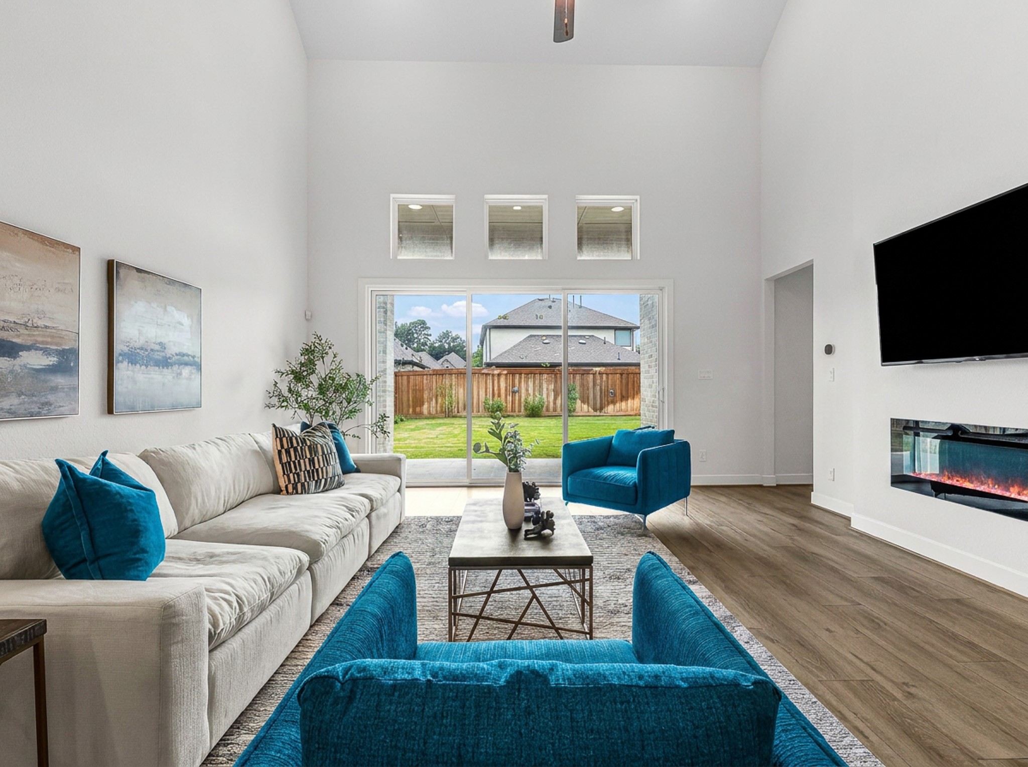 Soaring two-story ceilings and a sleek linear fireplace define this sunlit living area, where wide sliding glass doors open directly to the covered patio. Low-profile transom windows draw in additional light, complementing the warm wood-look floors and seamless access to the private green backyard.