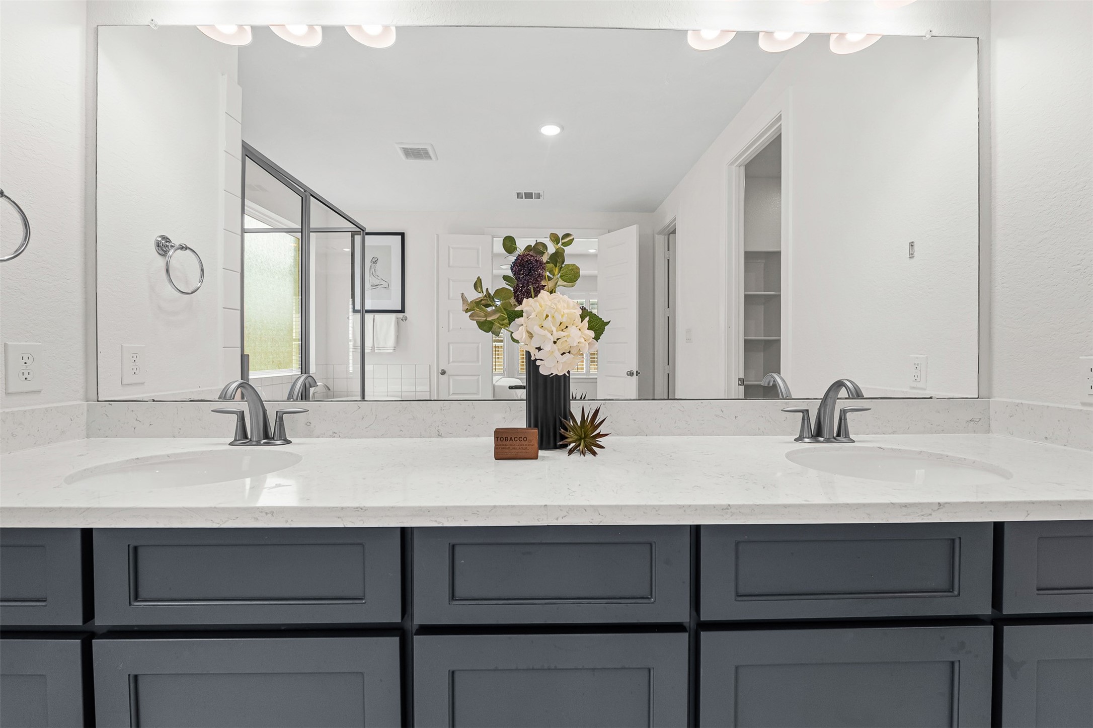 Dark contrast cabinetry and sleek white quartz countertops highlight this primary ensuite dual-sink vanity with brushed nickel fixtures. An expansive wall-to-wall mirror reflects the bright, spa-like atmosphere, including the glass-enclosed shower and freestanding soaking tub across the room.