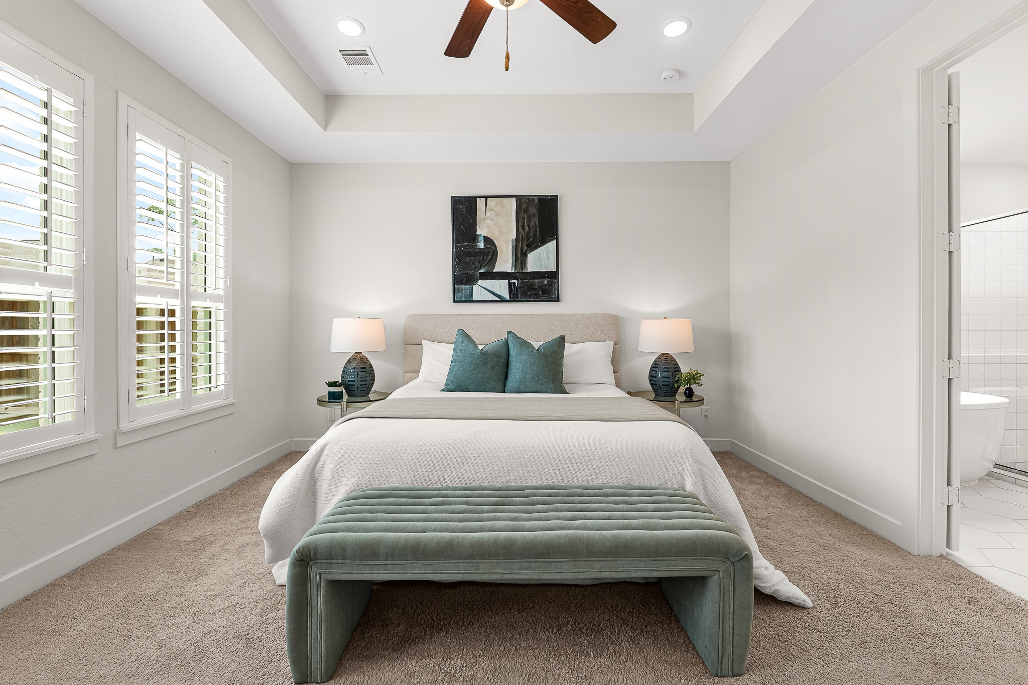 An elegant tray ceiling with a stylish fan crowns this first-floor primary suite, where soft neutral carpeting adds warmth to the expansive layout. White plantation shutters frame bright windows, and direct access to the luxurious ensuite bath completes the serene retreat.