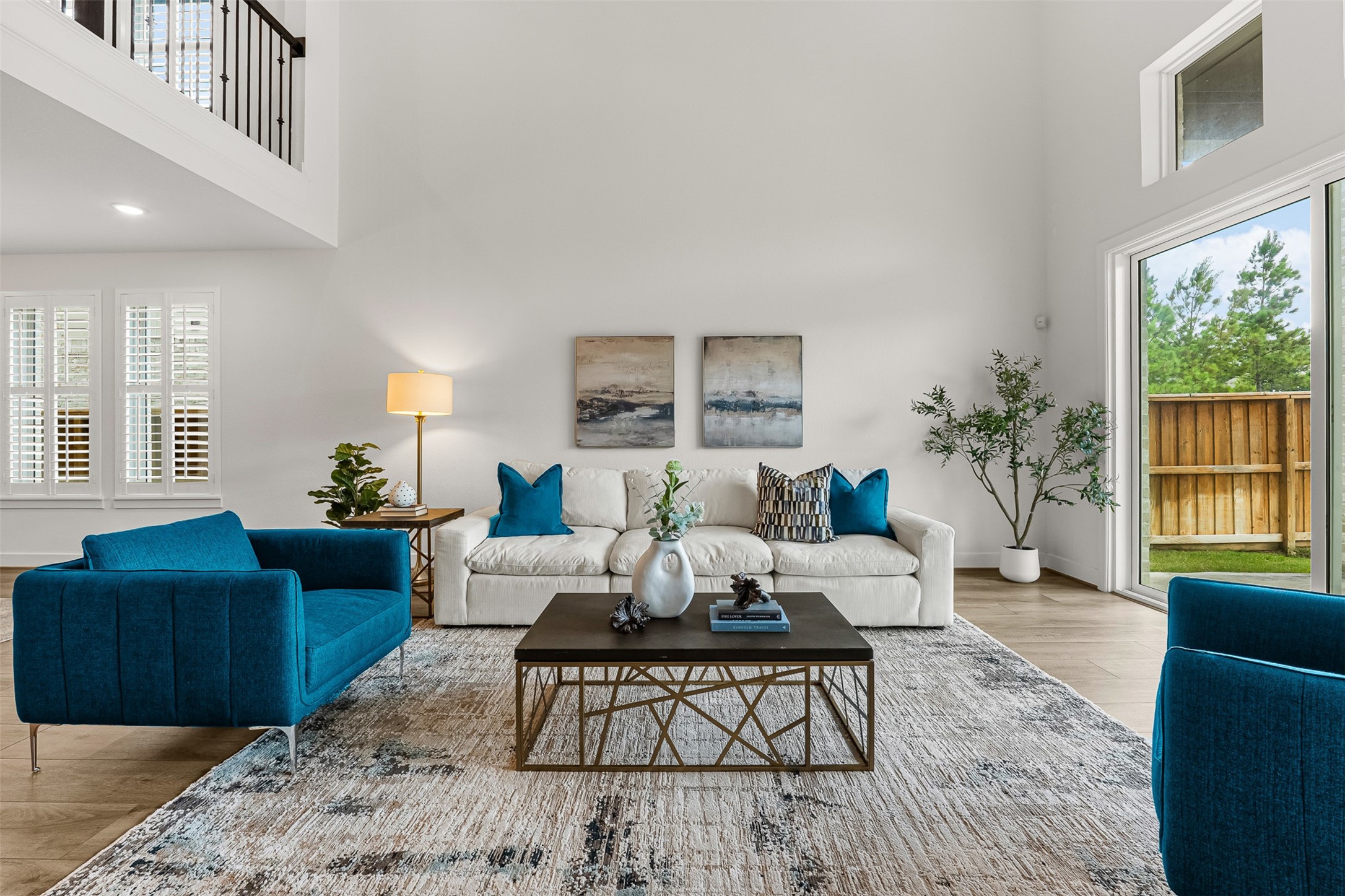 Soaring two-story ceilings and a second-floor catwalk overlook highlight this bright, open-concept first-floor living space. Sliding glass doors frame views of the private fenced yard, while planter shutters add polished privacy to the airy room.