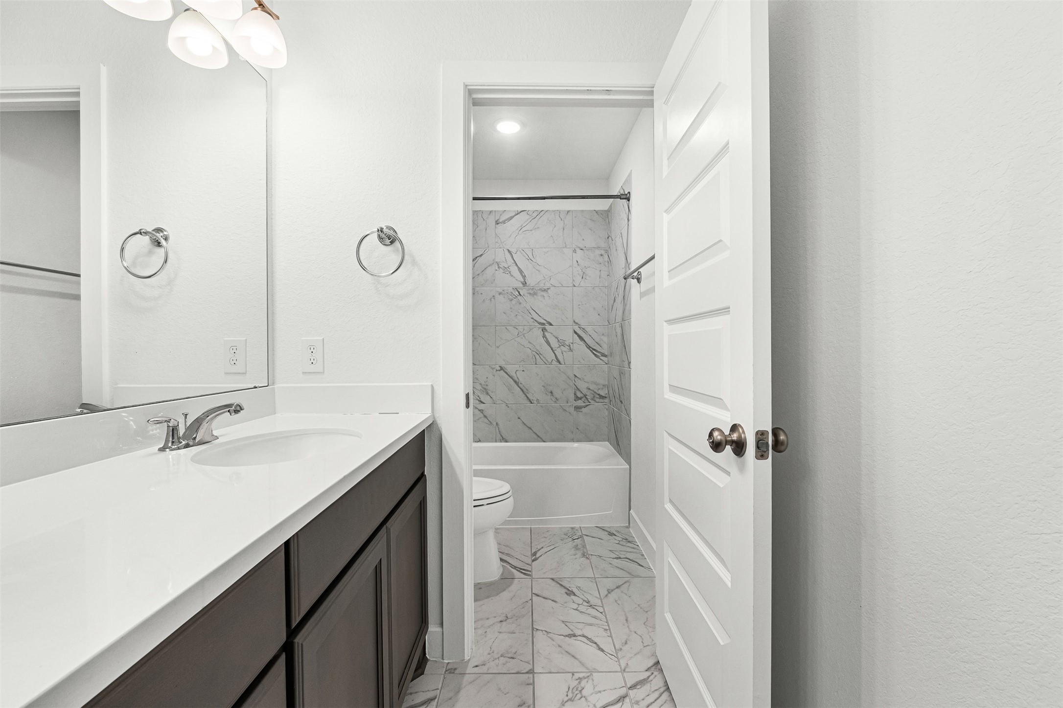 Refined marble-look tile flooring connects the spacious vanity area to a private water closet and tub-and-shower combination. A dark wood-look vanity with crisp white quartz countertops and sleek chrome fixtures adds modern function to this full bathroom.