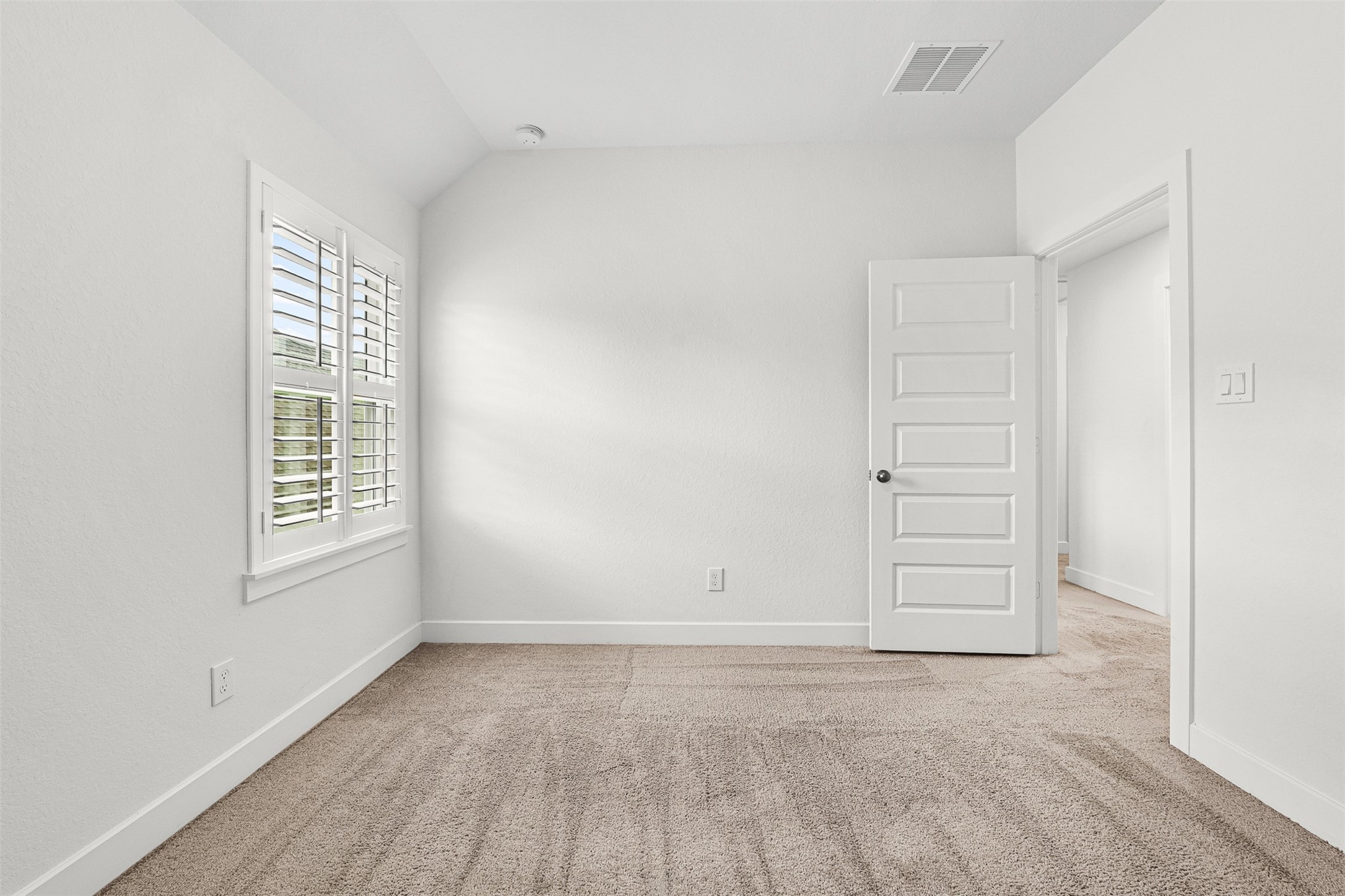 Clean plantation shutters frame the window in this secondary bedroom, featuring soft carpet flooring and classic neutral walls. A versatile layout and sloped ceiling details provide a peaceful space for rest or personalized furnishings.