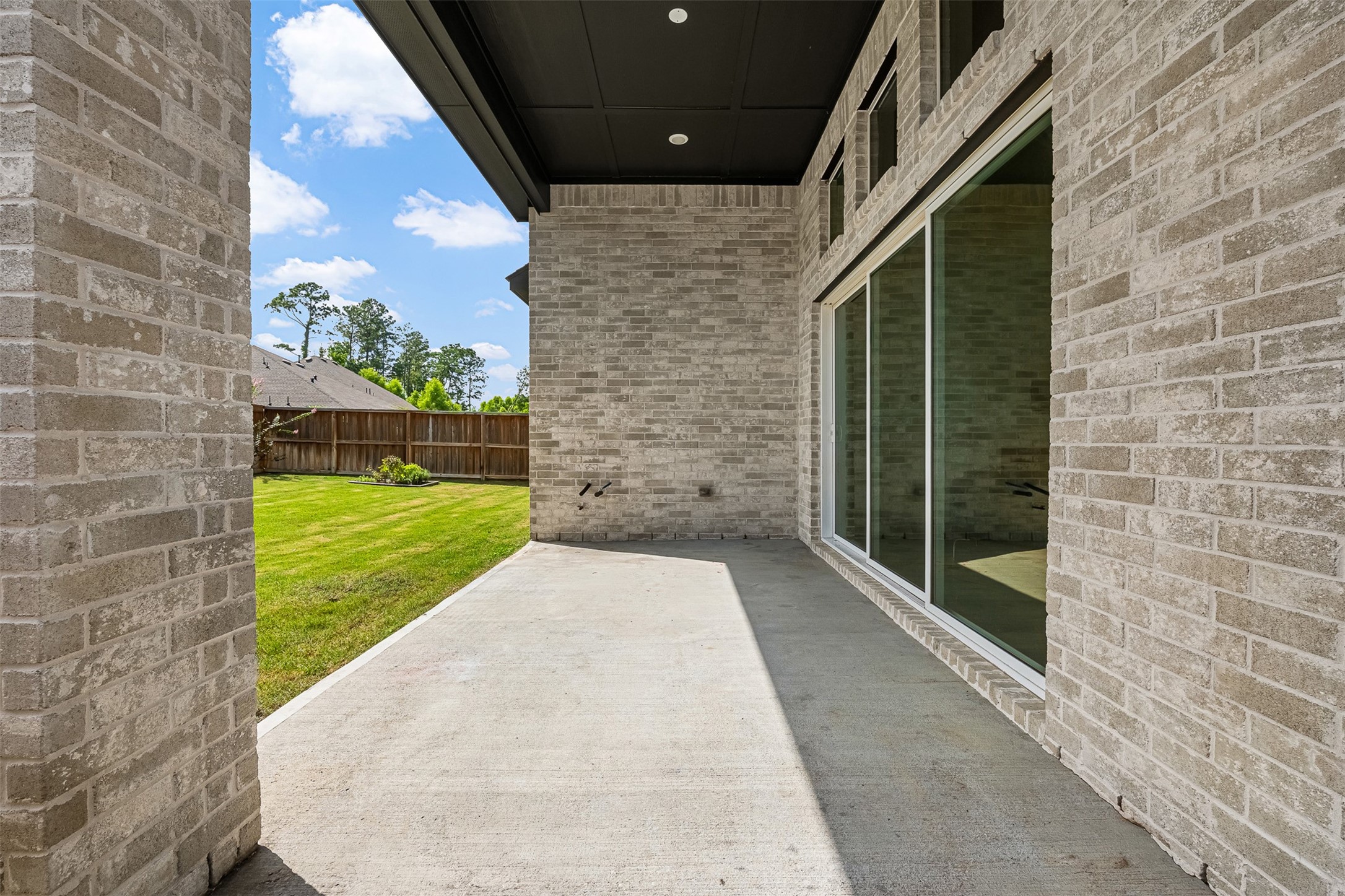 Extending from the home's rear exterior, this covered concrete patio provides an inviting shelter for outdoor relaxation. Soaring brick walls and dark tongue-and-groove ceiling accents frame the space, offering direct access to the lush green lawn and expansive sliding glass doors.