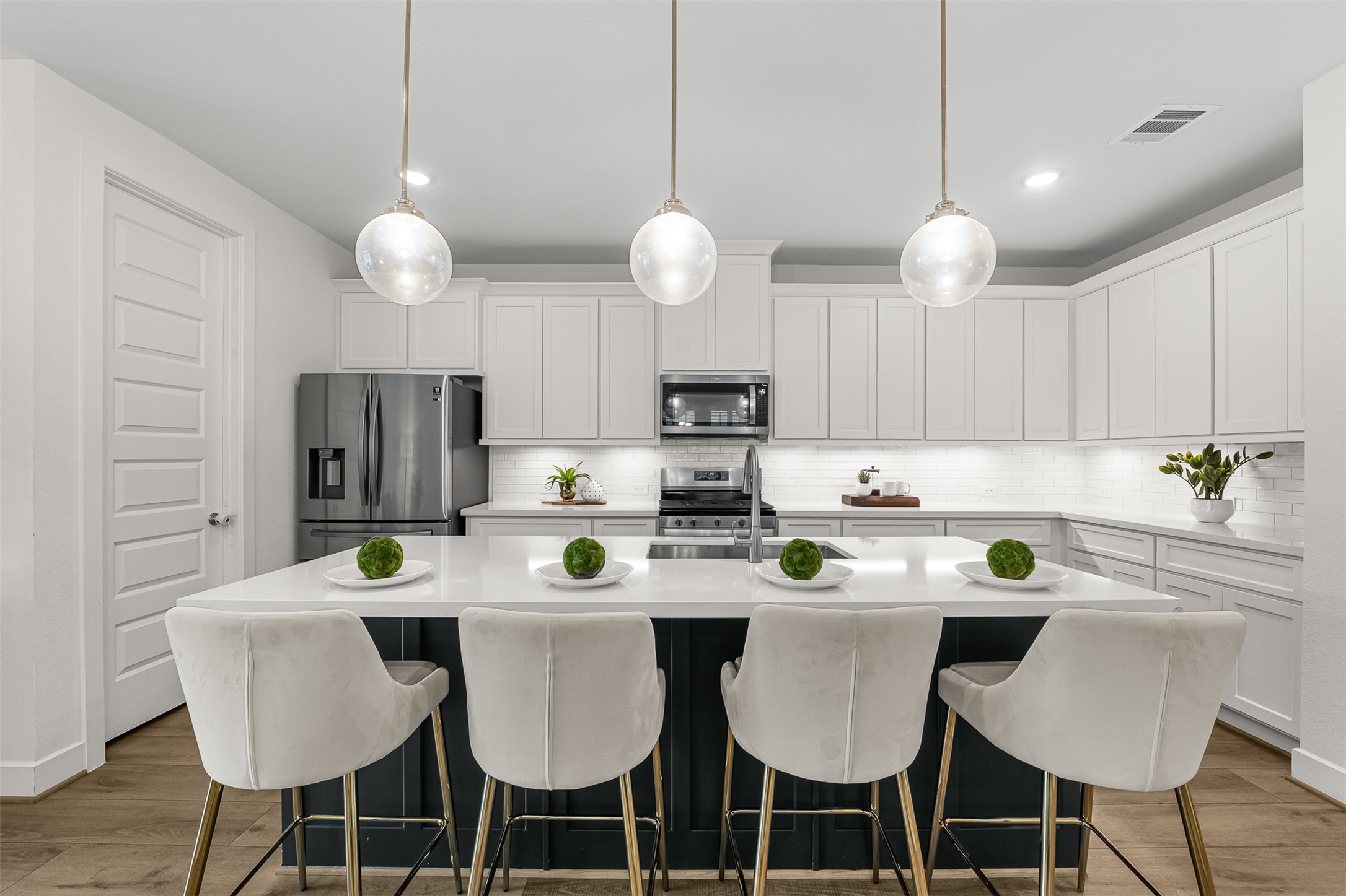 Sleek white cabinetry and crisp tile backsplash surround stainless steel appliances, creating a polished environment with a spacious walk-in pantry nearby. Under-cabinet lighting illuminates expansive quartz counter space, while a central sink integrates seamlessly into the contrasting island.