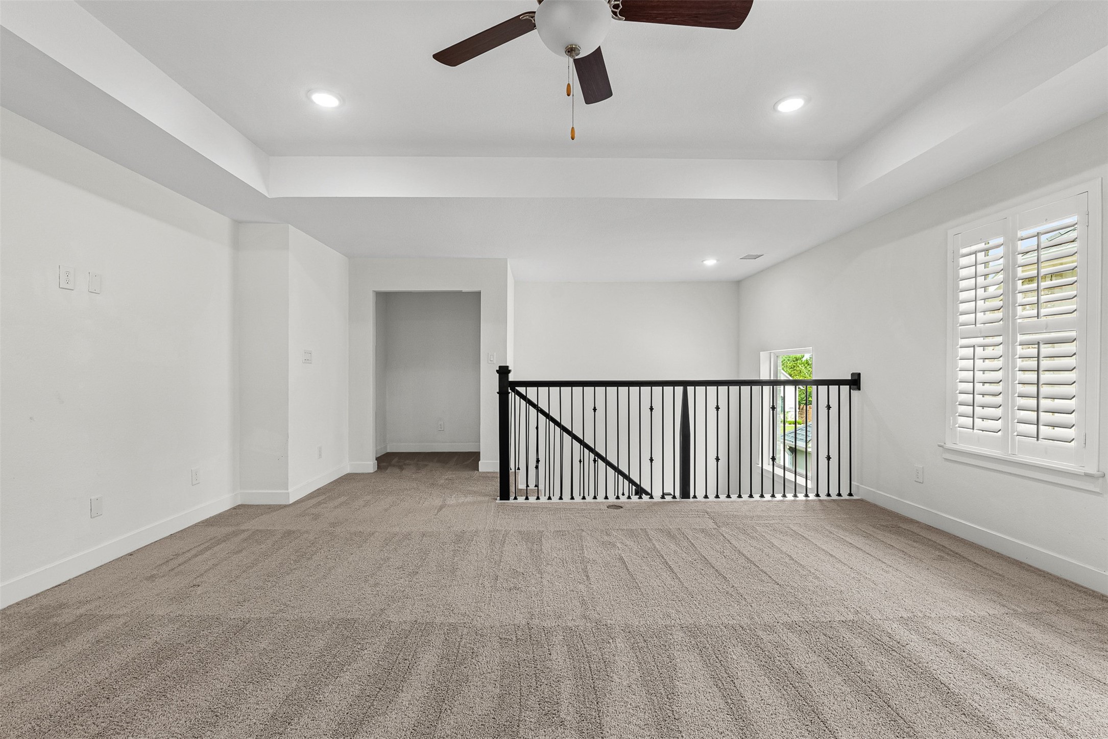 Wrought-iron railings overlook the lower level from this expansive second-floor game room featuring a tray ceiling and plush carpet. Plantation shutters frame the window, while a recessed alcove provides a quiet nook for desk placement or extra storage.