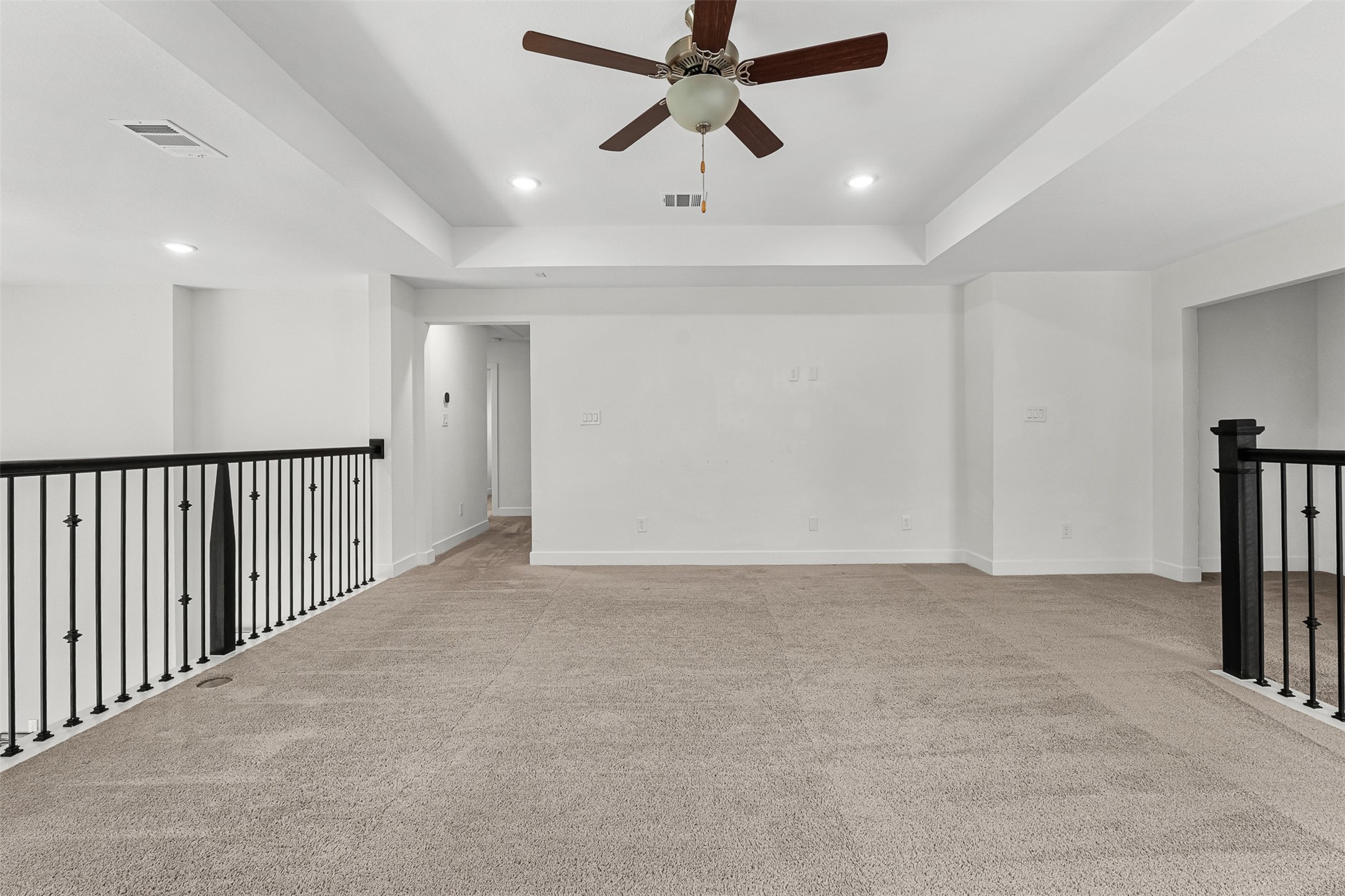 Perched above the first floor, this versatile flex space features plush carpet, a tray ceiling, and a dark wrought-iron railing overlook. Recessed lighting illuminates the open layout, while a central doorway leads to adjoining secondary bedrooms.