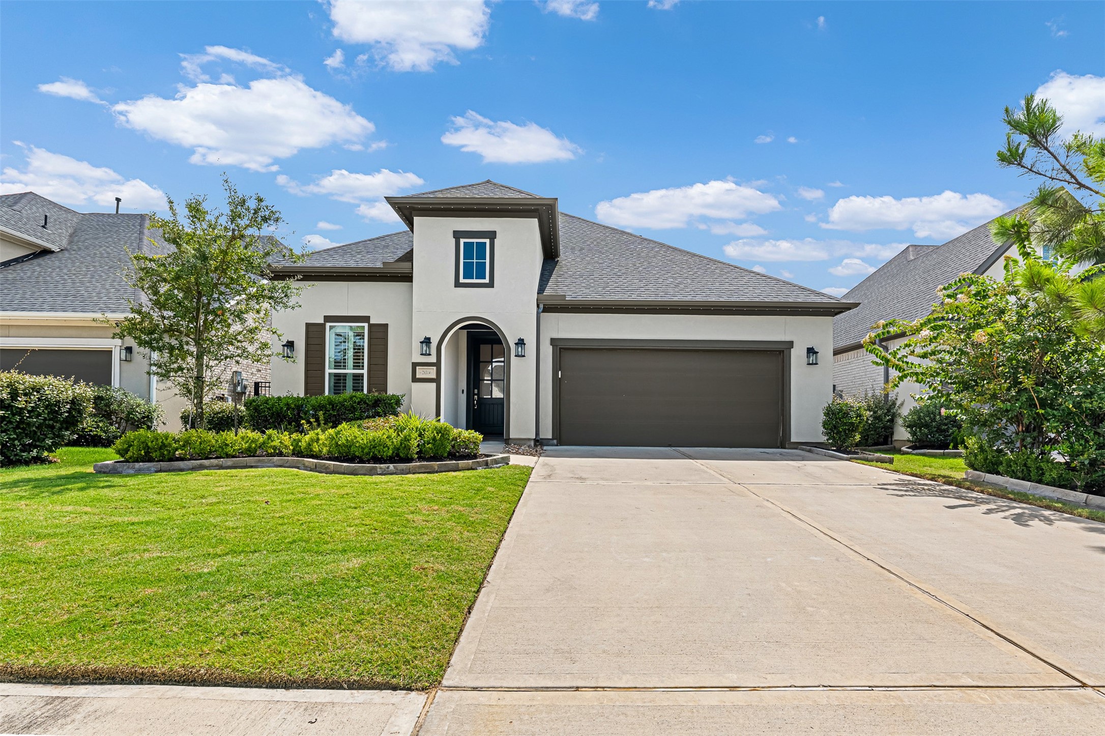Featuring a sleek stucco finish, dark accent shutters, and an arched entry, this two-story contemporary home makes a striking impression. An expansive concrete driveway leads to the attached two-car garage, framed by manicured green space and stone-bordered landscaping.