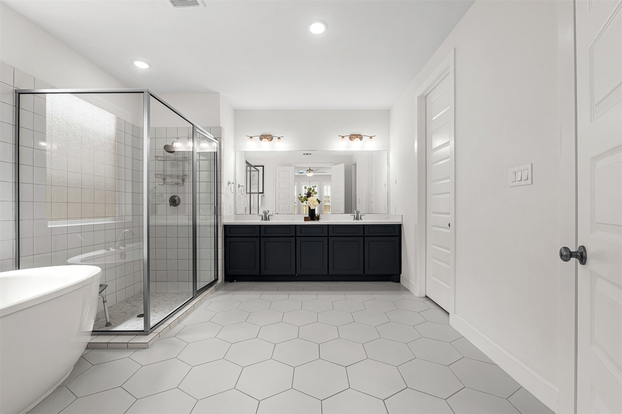 Large-format hexagon tile floors and crisp white walls frame this spa-like primary bath, complete with a freestanding soaking tub and glass-enclosed walk-in shower. A dark contrast vanity with dual sinks, quartz countertops, and classic multi-light sconces provides a striking centerpiece.