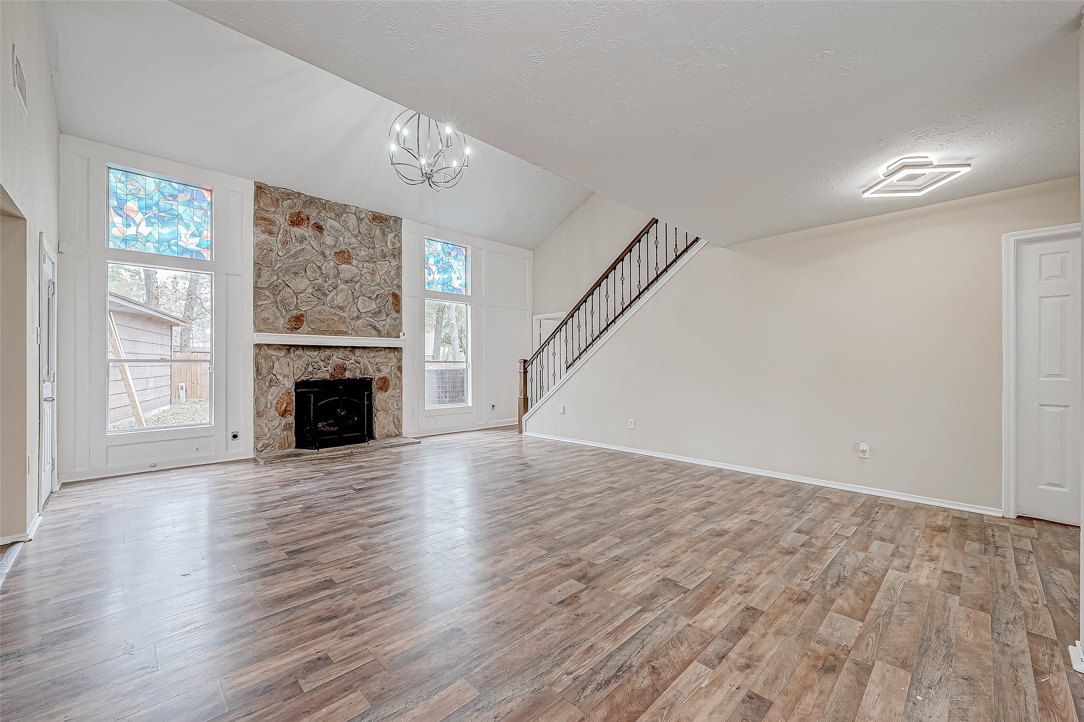 Anchored by a striking fireplace that reaches toward the soaring ceiling, this living room exudes grandeur. Large windows, including elegant stained-glass panels, bathe the room in natural light while offering a touch of artistry. Expansive vinyl flooring complements the open layout, creating a dynamic space.