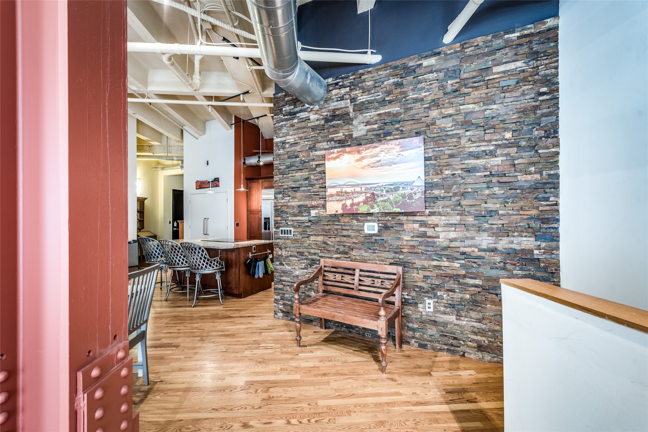 This space features a modern industrial design with exposed ductwork and a stone accent wall. The room includes a cozy bench and leads into a kitchen area with a breakfast bar and stylish seating. Warm wood flooring enhances the inviting atmosphere.