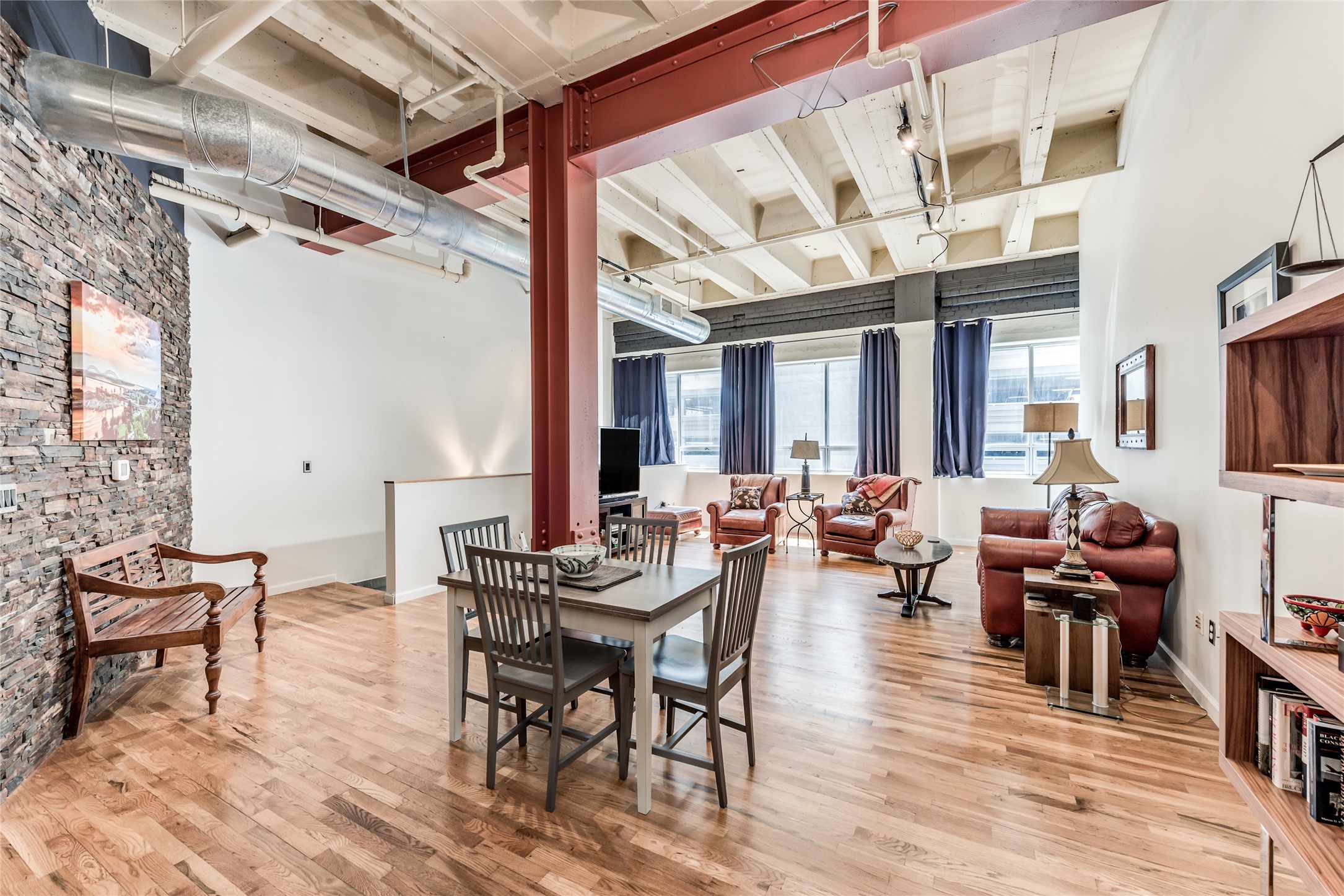 This photo showcases a spacious, open-concept loft with high ceilings, exposed ductwork, and a mix of industrial and cozy elements. The room features polished wood floors, a brick accent wall, and large windows with ample natural light. The living area includes comfortable seating and a dining space, perfect for modern urban living. This photo showcases a spacious, open-concept loft with high ceilings, exposed ductwork, and a mix of industrial and cozy elements. The room features polished wood floors, a brick accent wall, and large windows with ample natural light. The living area includes comfortable seating and a dining space, perfect for modern urban living.