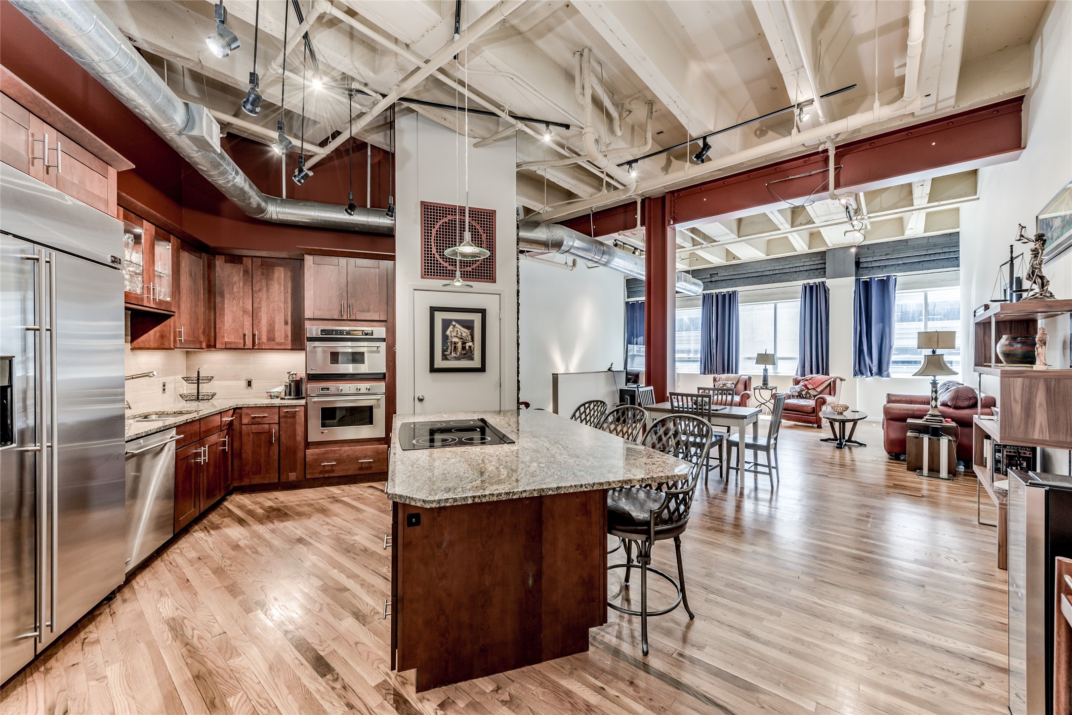This photo showcases a spacious, modern open-concept living area with a sleek kitchen featuring stainless steel appliances, a central island with cook top and breakfast bar seating, and rustic wood cabinetry. The space extends into a light-filled living and dining area, highlighting high ceilings with exposed beams and ductwork, creating an industrial yet inviting atmosphere. This photo showcases a spacious, modern open-concept living area with a sleek kitchen featuring stainless steel appliances, a central island with cook top and breakfast bar seating, and rustic wood cabinetry. The space extends into a light-filled living and dining area, highlighting high ceilings with exposed beams and ductwork, creating an industrial yet inviting atmosphere.