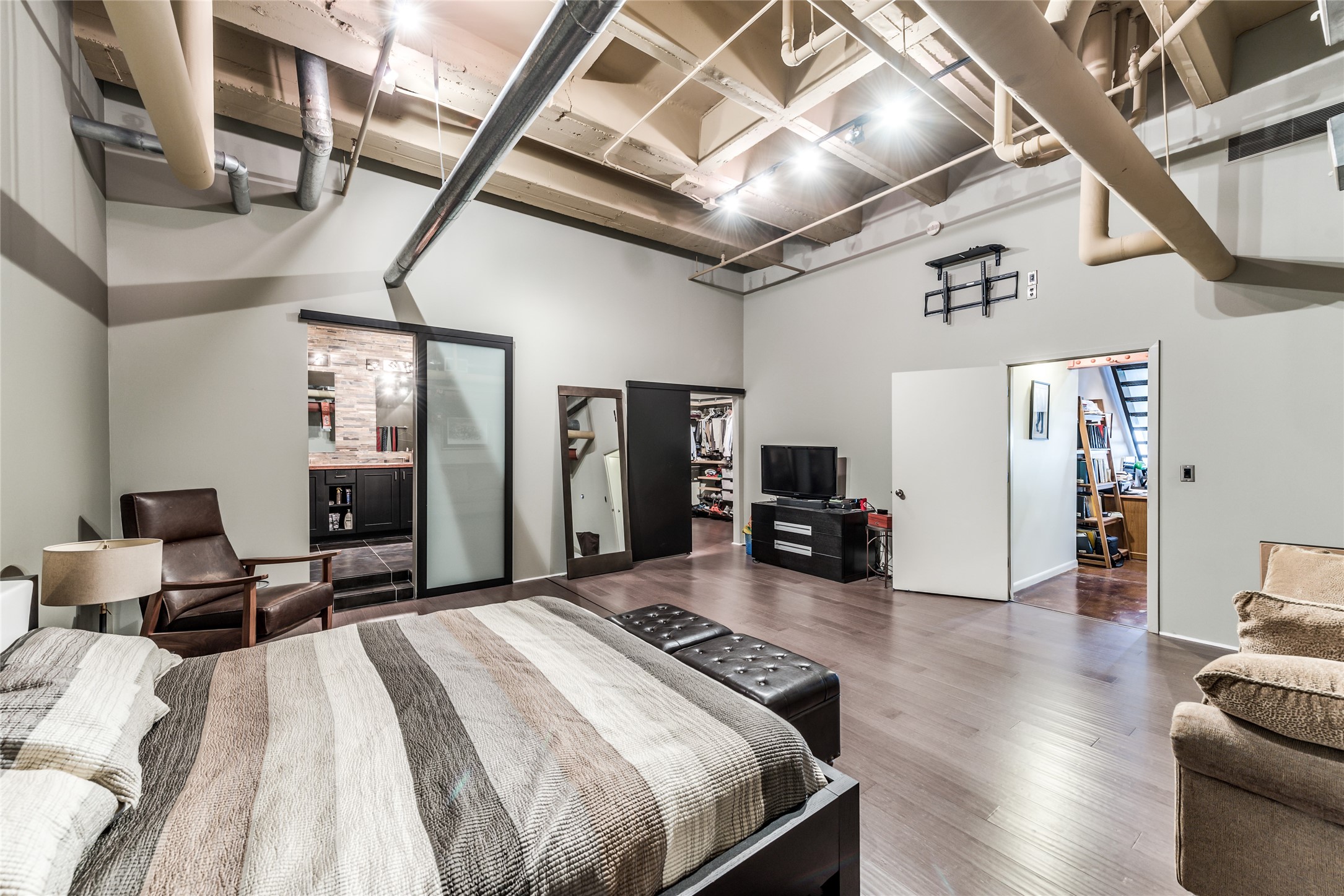 This spacious, industrial-style primary bedroom features high ceilings with exposed ductwork, modern lighting, and wood flooring.