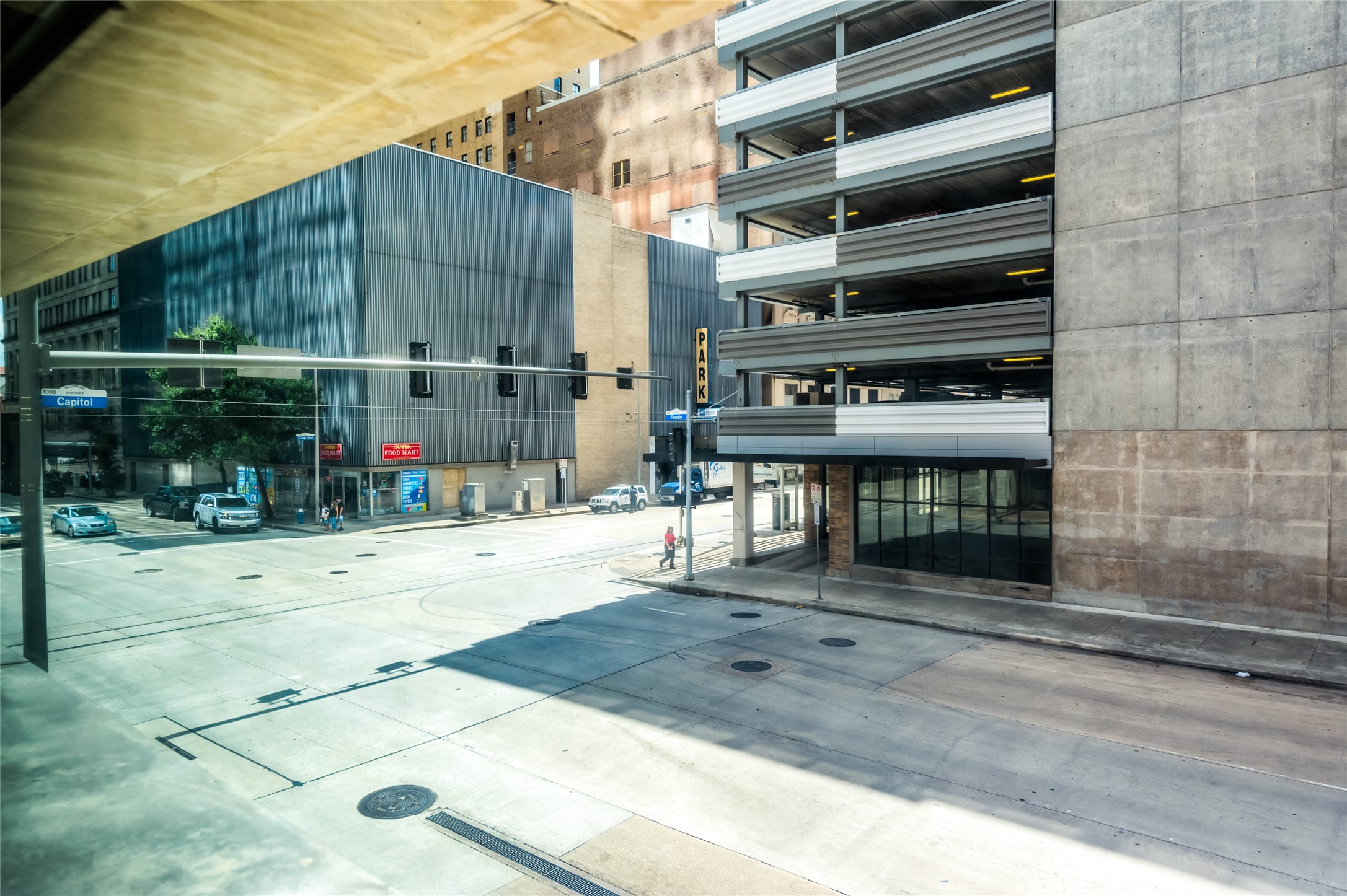 Urban view featuring a street corner with a parking garage and modern commercial buildings. Wide sidewalks and minimal traffic provide a spacious city feel.