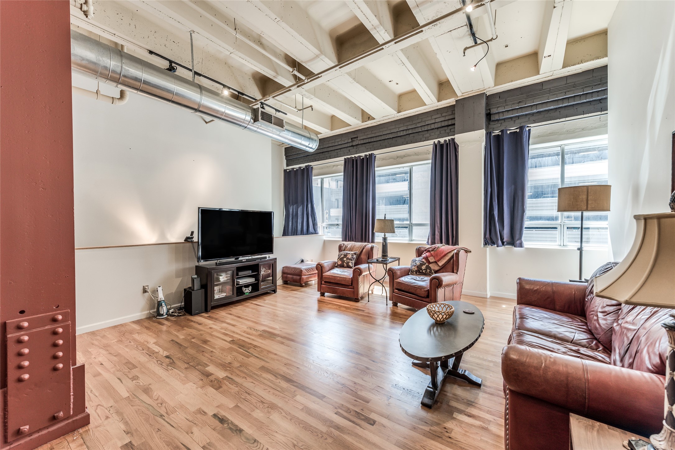 This photo showcases a spacious, industrial-style living room with high ceilings, exposed beams, and ductwork. It features large windows, hardwood floors, leather furniture, and a cozy seating area with a TV. The space is bright and modern, ideal for comfortable living. This photo showcases a spacious, industrial-style living room with high ceilings, exposed beams, and ductwork. It features large windows, hardwood floors, leather furniture, and a cozy seating area with a TV. The space is bright and modern, ideal for comfortable living.