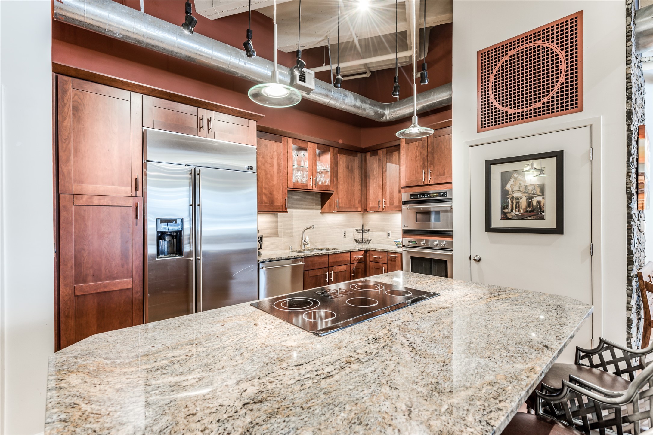 Modern kitchen with stainless steel appliances, granite countertops, and rich wooden cabinetry. Features an island with a built-in cooktop and industrial-style lighting, offering a stylish and functional space. Modern kitchen with stainless steel appliances, granite countertops, and rich wooden cabinetry. Features an island with a built-in cooktop and industrial-style lighting, offering a stylish and functional space.