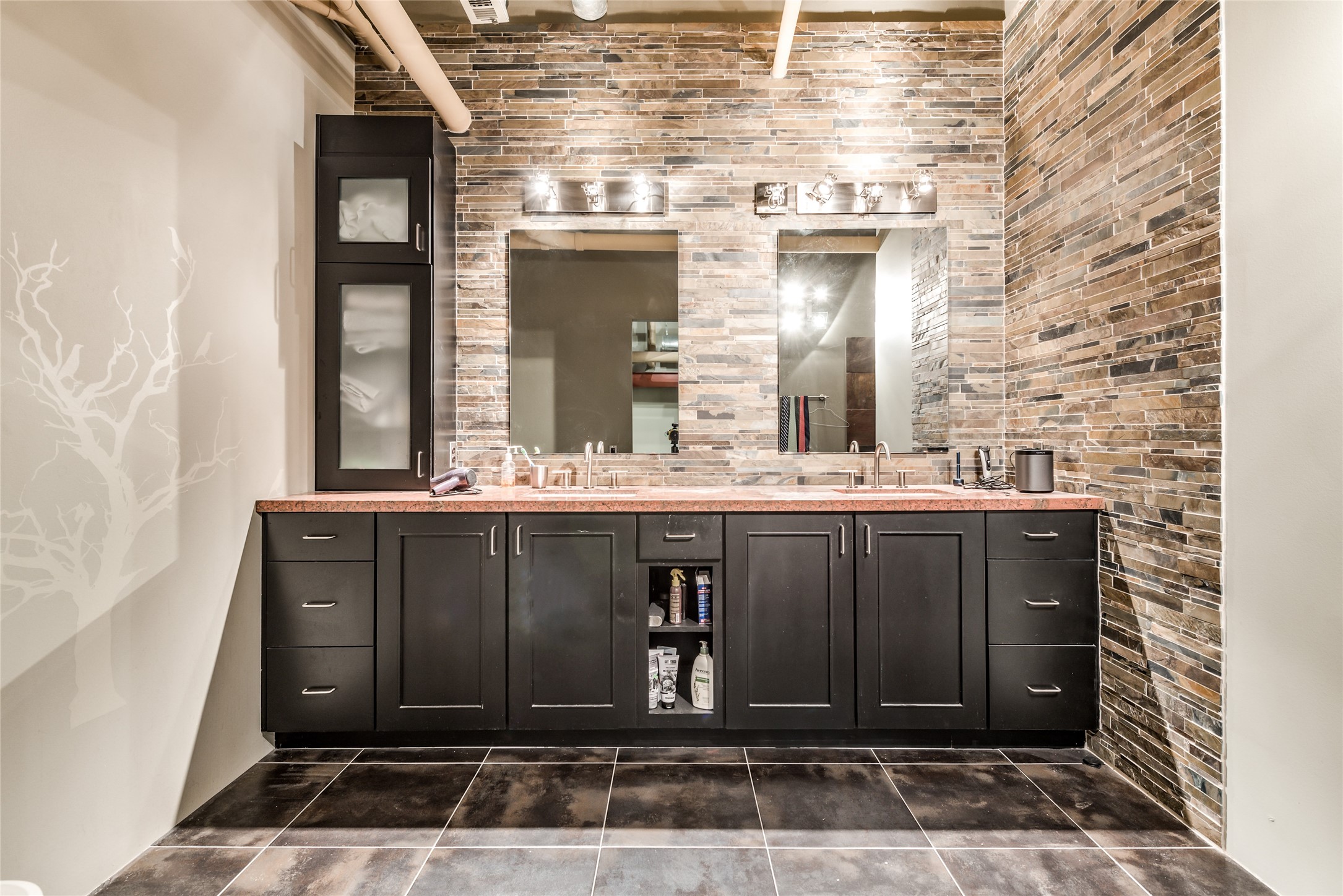 This modern primary bathroom features a double vanity with dark cabinetry and a sleek stone-tiled wall.