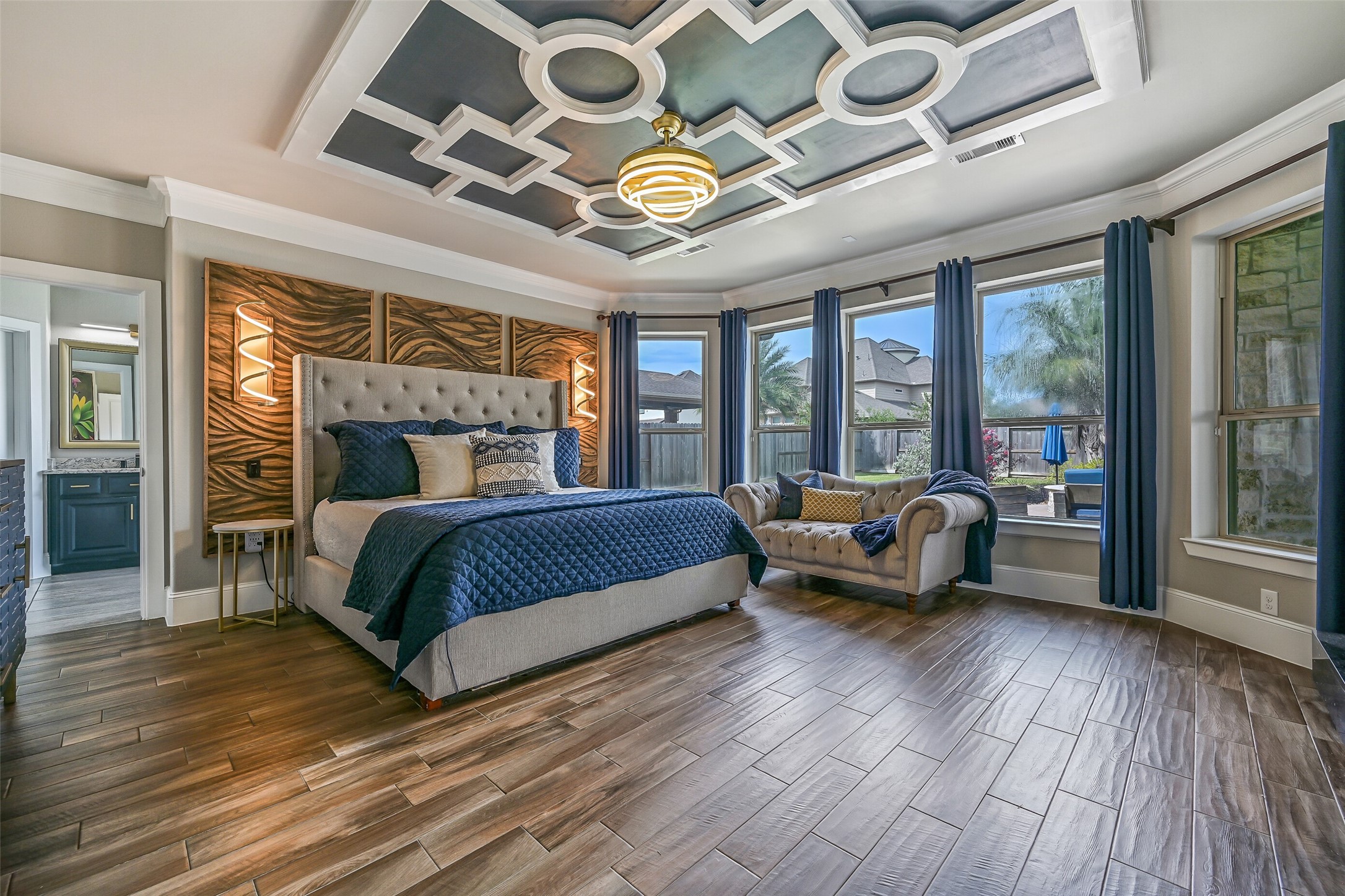 The secluded first-floor primary retreat is filled with custom detail, from the dramatic geometric ceiling treatment, modern lighting, & linear fireplace to wood-look tile floors and a wall of windows overlooking the backyard pool.
