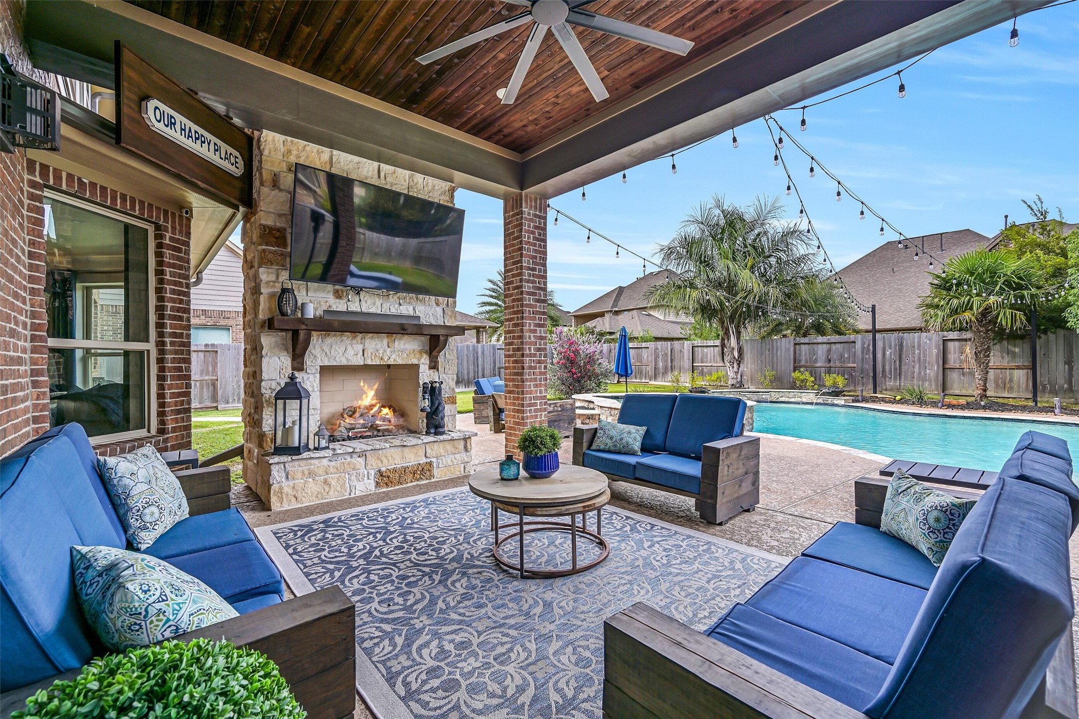 This one may be my favorite patio shot! The covered outdoor living room centers around a handsome Austin limestone gas-log fireplace with mounted TV, beneath a stained wood ceiling with oversized fan and pool views beyond.