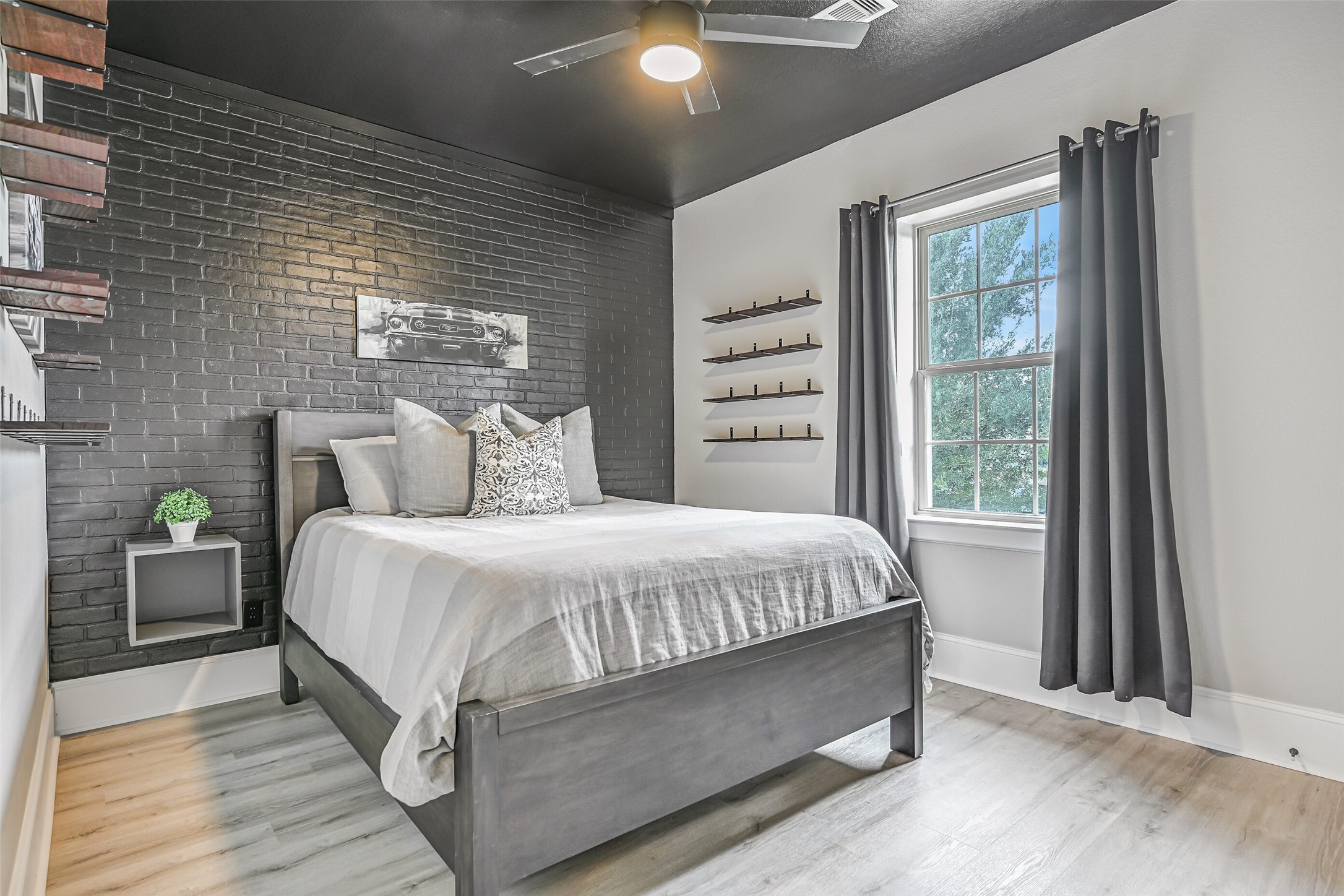 This upstairs secondary bedroom features LVP flooring, a dramatic black-painted brick accent wall, black ceiling, lighted ceiling fan and industrial-style shelving. A walk-in closet and Jack & Jill bath connect it to the adjoining bedroom.