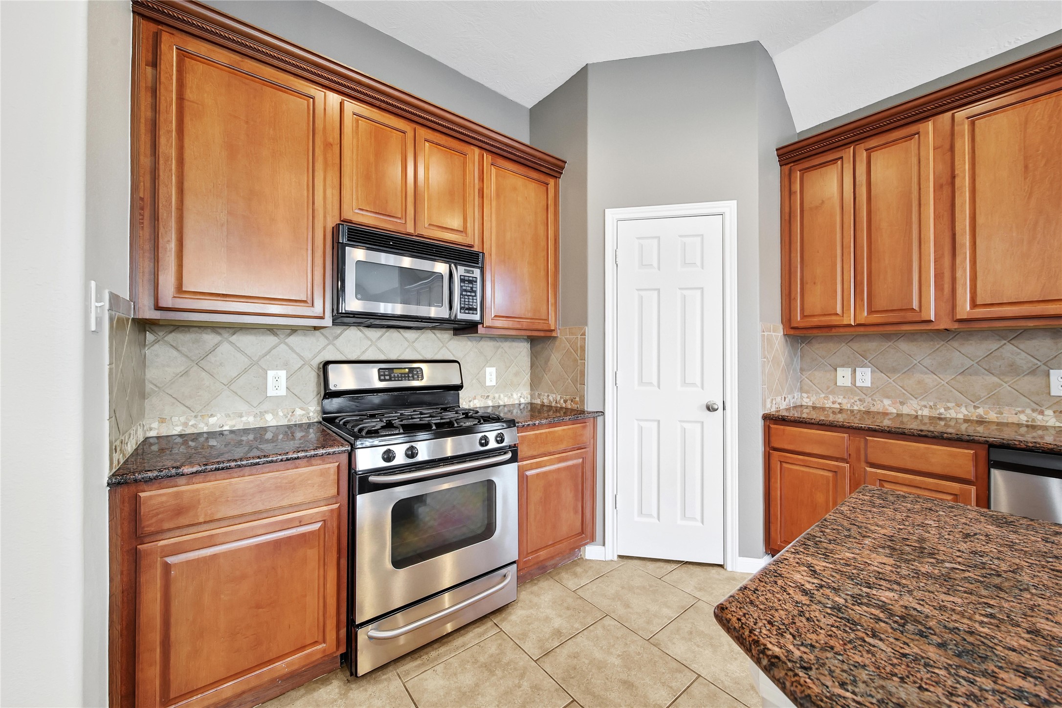 This kitchen features warm-toned wooden cabinetry, granite countertops, stainless steel appliances, a built-in microwave, and a gas stove. It has tiled backsplash and flooring, with a neutral color scheme.