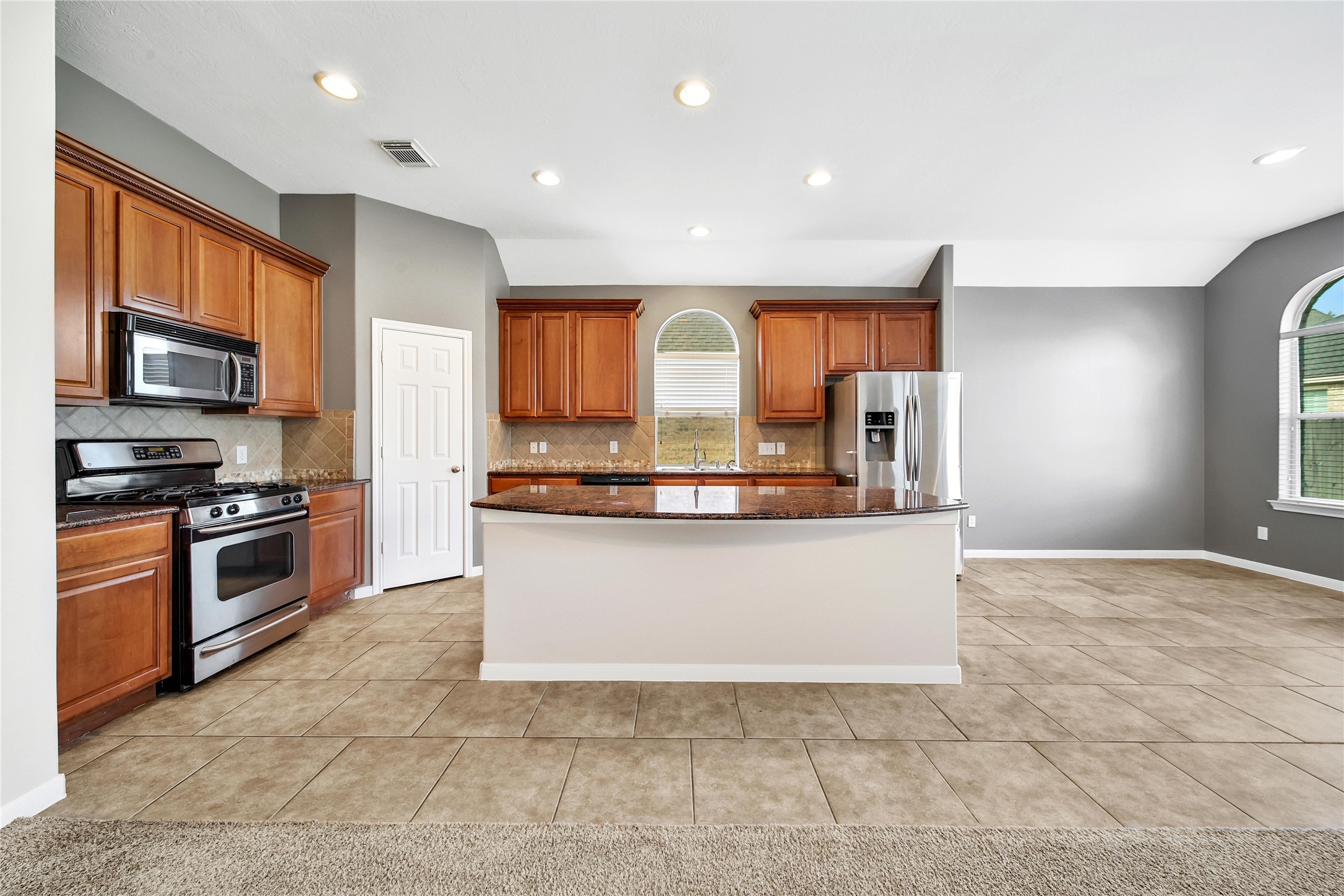 This is a spacious kitchen featuring stainless steel appliances, ample wooden cabinetry, a central island with a countertop, and a mix of tile and carpeted flooring. The area is well-lit with natural light from large windows and has recessed ceiling lights.
