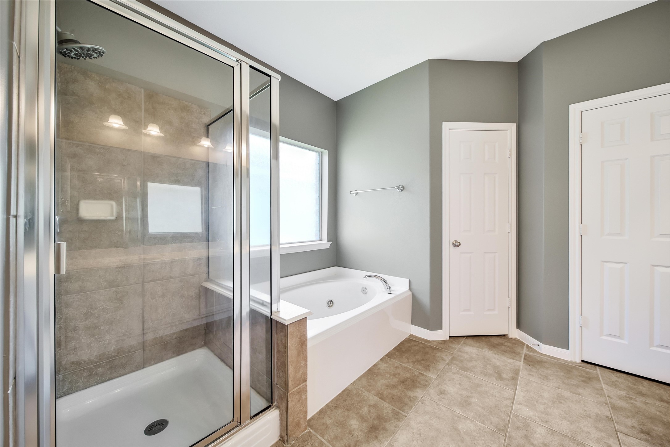 The modern looking primary bathroom features an oversized glass-enclosed shower, a separate bathtub, tiled flooring, and neutral wall colors. It includes a window for natural light, and a huge closet.