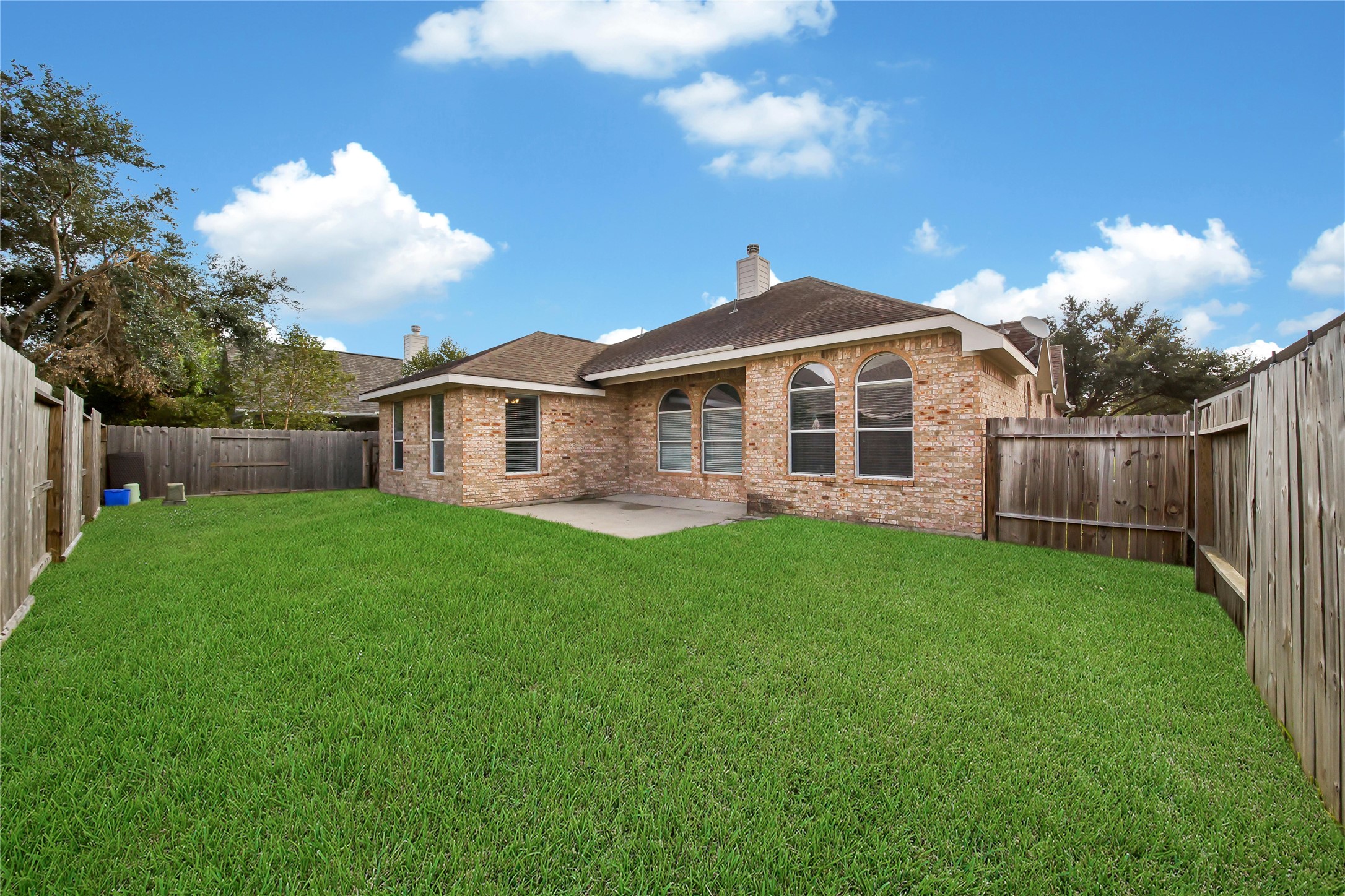 This image showcases a single-story brick home featuring a lawn and a fenced backyard, with ample outdoor space for activities and relaxation. The house has large windows for natural lighting and a concrete patio area.