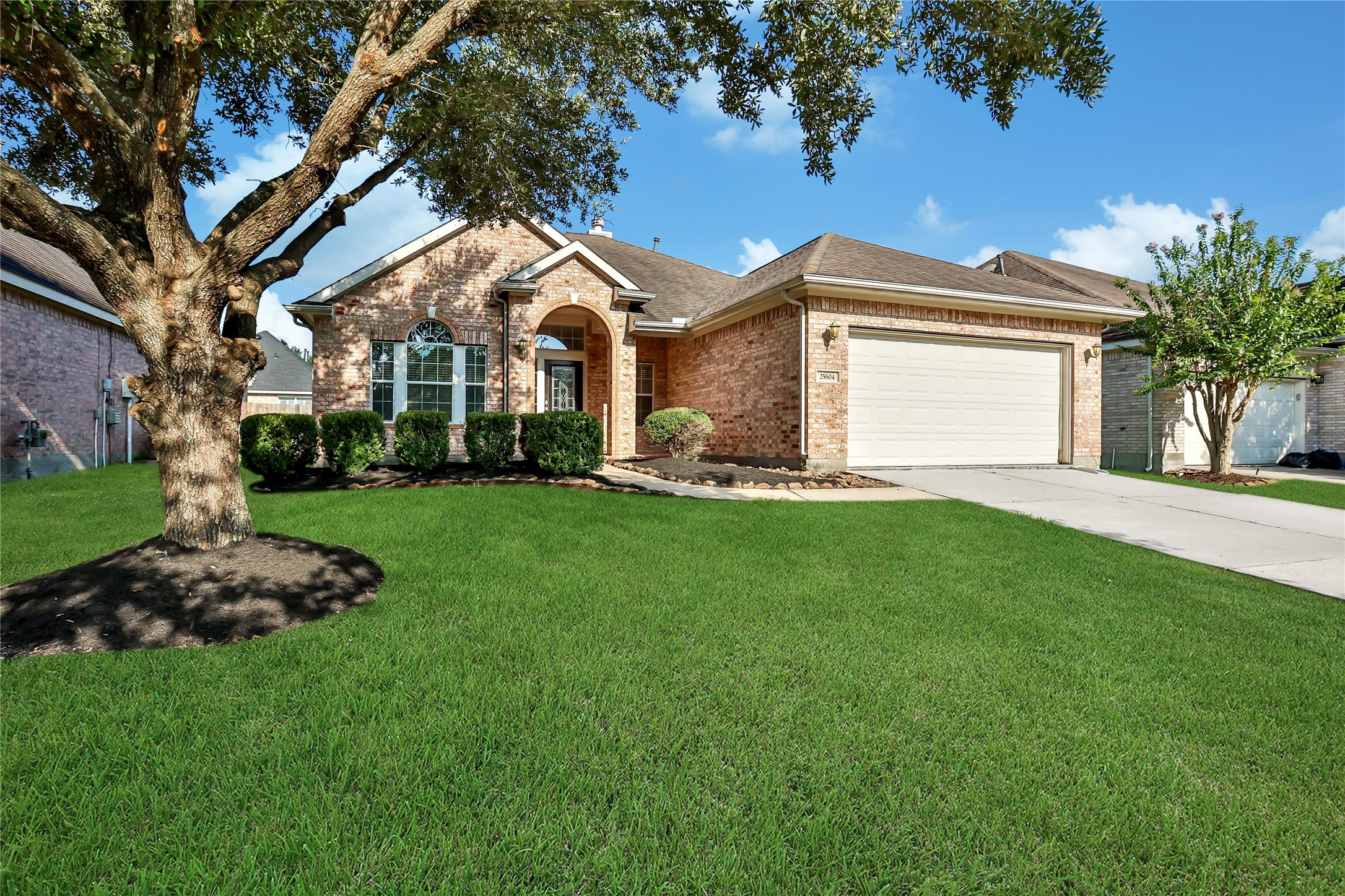 This is a well-maintained single-story brick home featuring a two-car garage, a mature oak tree in the front yard, and neat landscaping.
