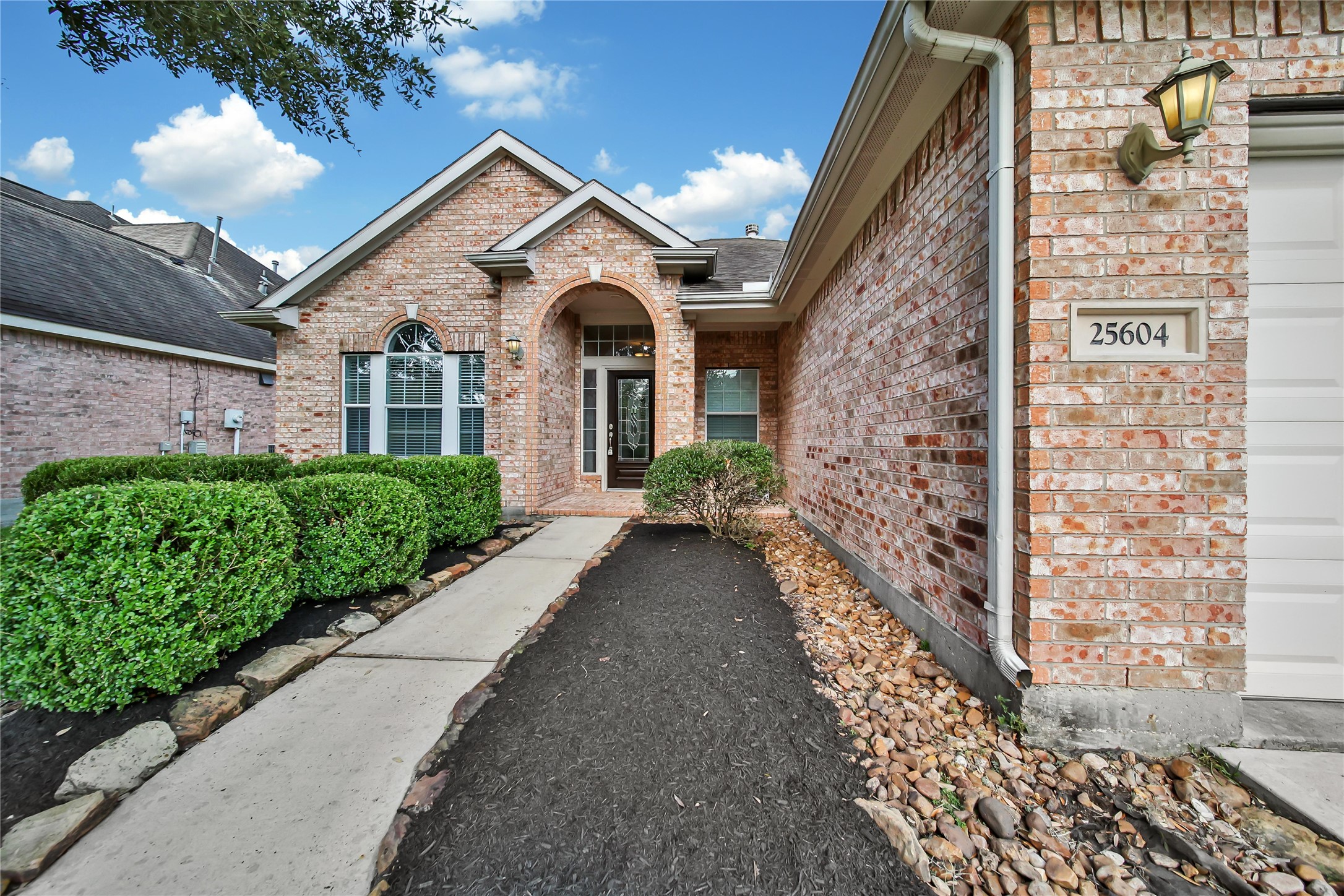 This is a single-story brick home featuring a well-maintained exterior with neat landscaping and a welcoming path leading to an arched entryway. The home has a pitched roof and a large front window, presenting a classic suburban appearance.
