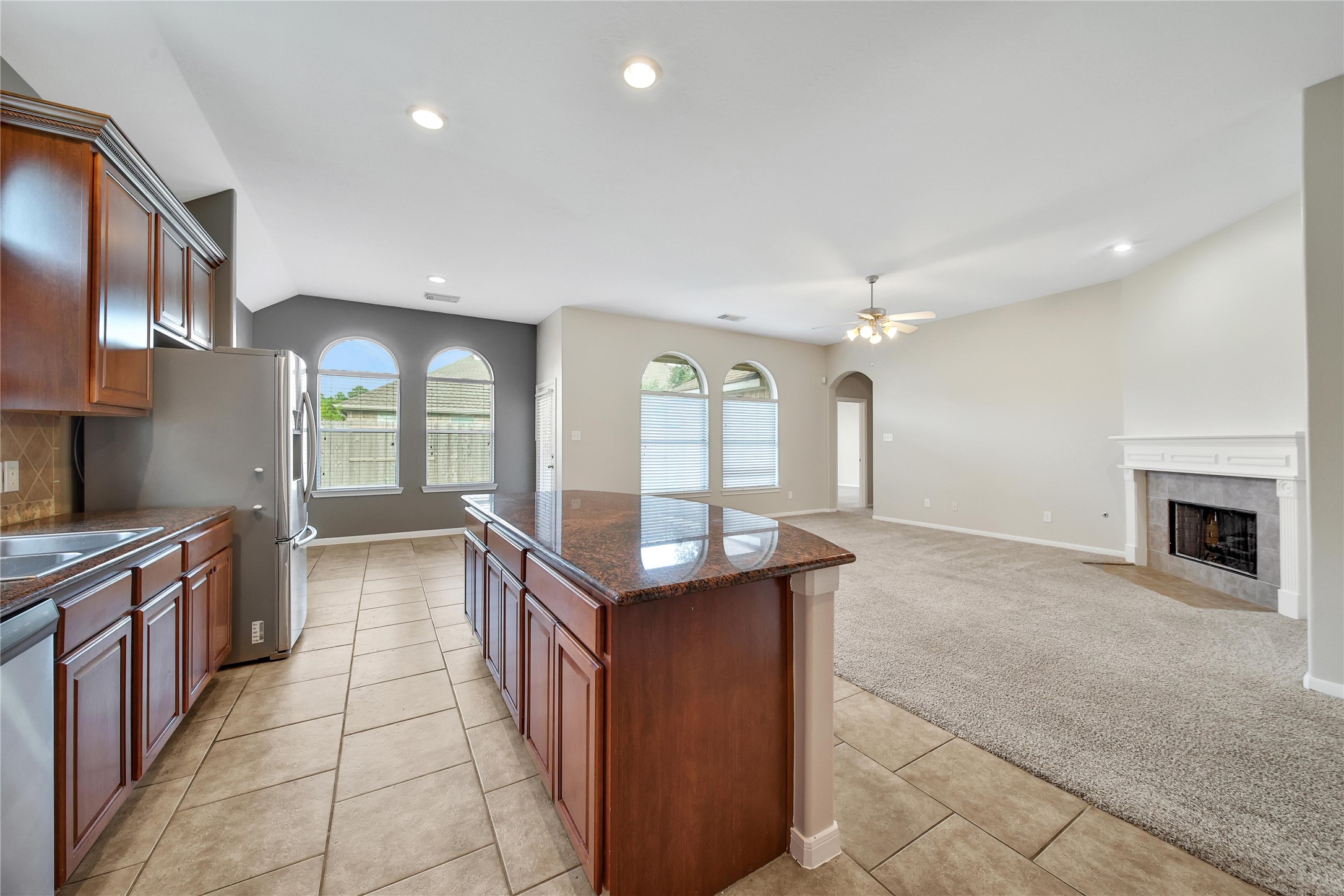 This is a spacious, open-plan living area with a modern kitchen featuring stainless steel appliances, wooden cabinets, and a central island. The room transitions into a cozy carpeted living space with ample natural light, a fireplace, and a view of the outdoors through large windows.