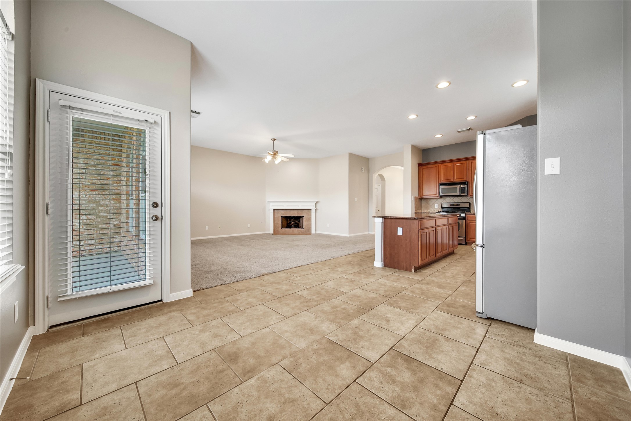 This is a spacious, open-concept living area with tile flooring, leading into a carpeted section with a cozy fireplace. The adjoining kitchen features wooden cabinets and modern appliances, with natural light streaming in from the glass door located off the breakfast room.