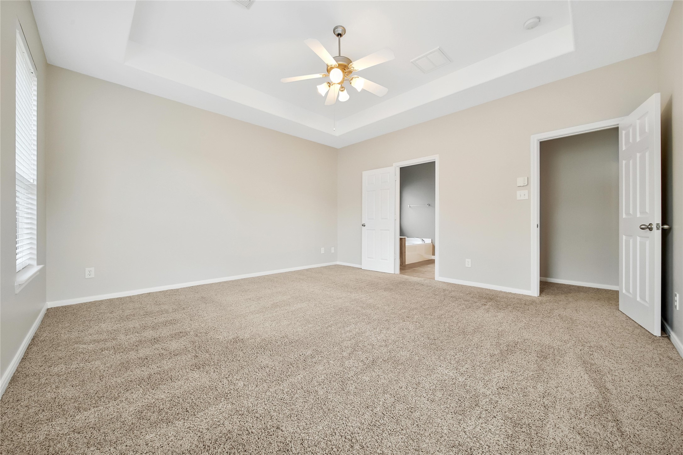 This is a spacious, well-lit room Primary Bedroom with neutral walls and carpet flooring, featuring a tray ceiling with a ceiling fan and double doors leading to the well appointed primary bath.