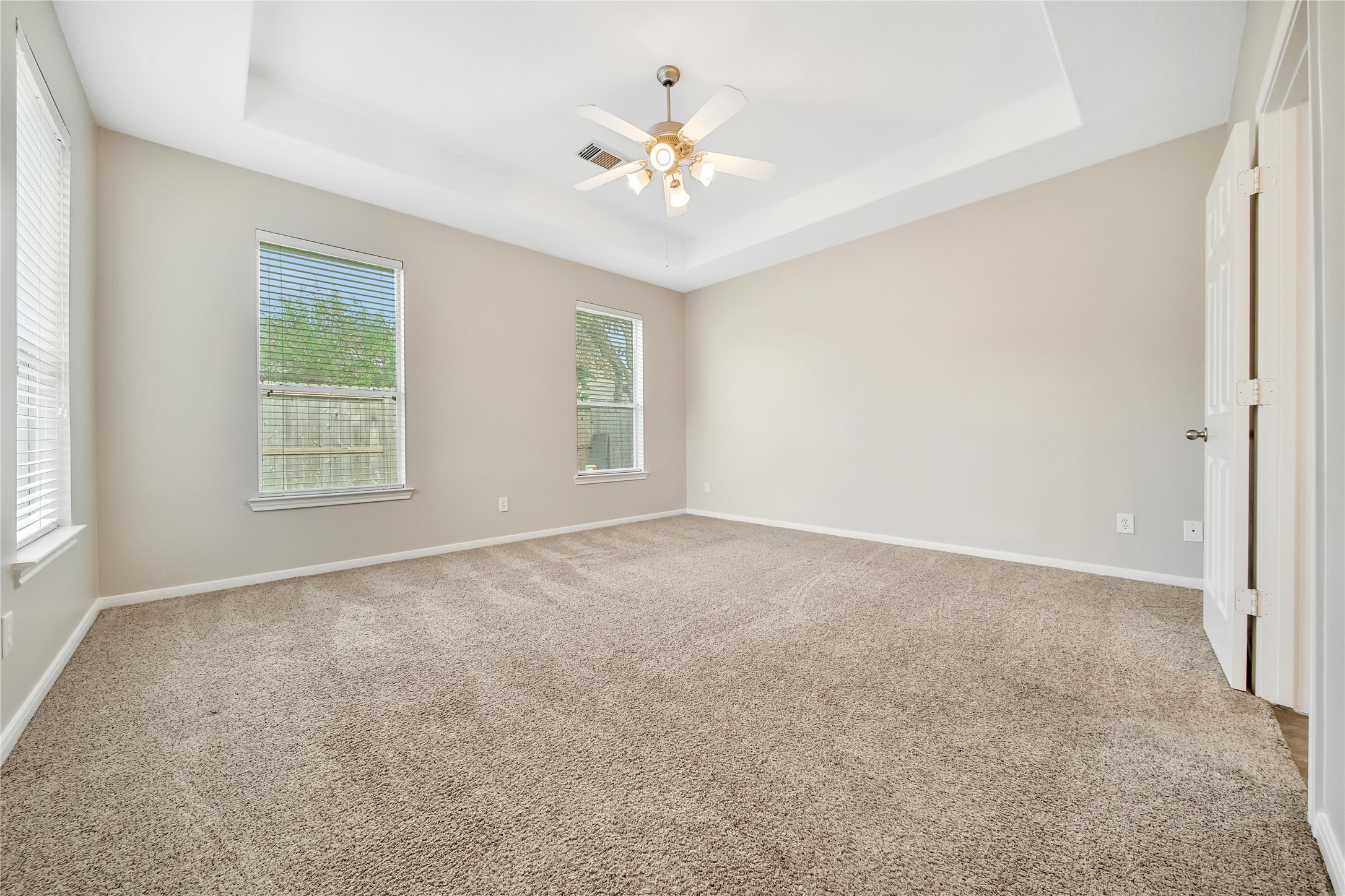 This is a spacious, well-lit Primary Bedroom with neutral walls, carpeting, and a ceiling fan with lighting. It features two large windows that allow for ample natural light.